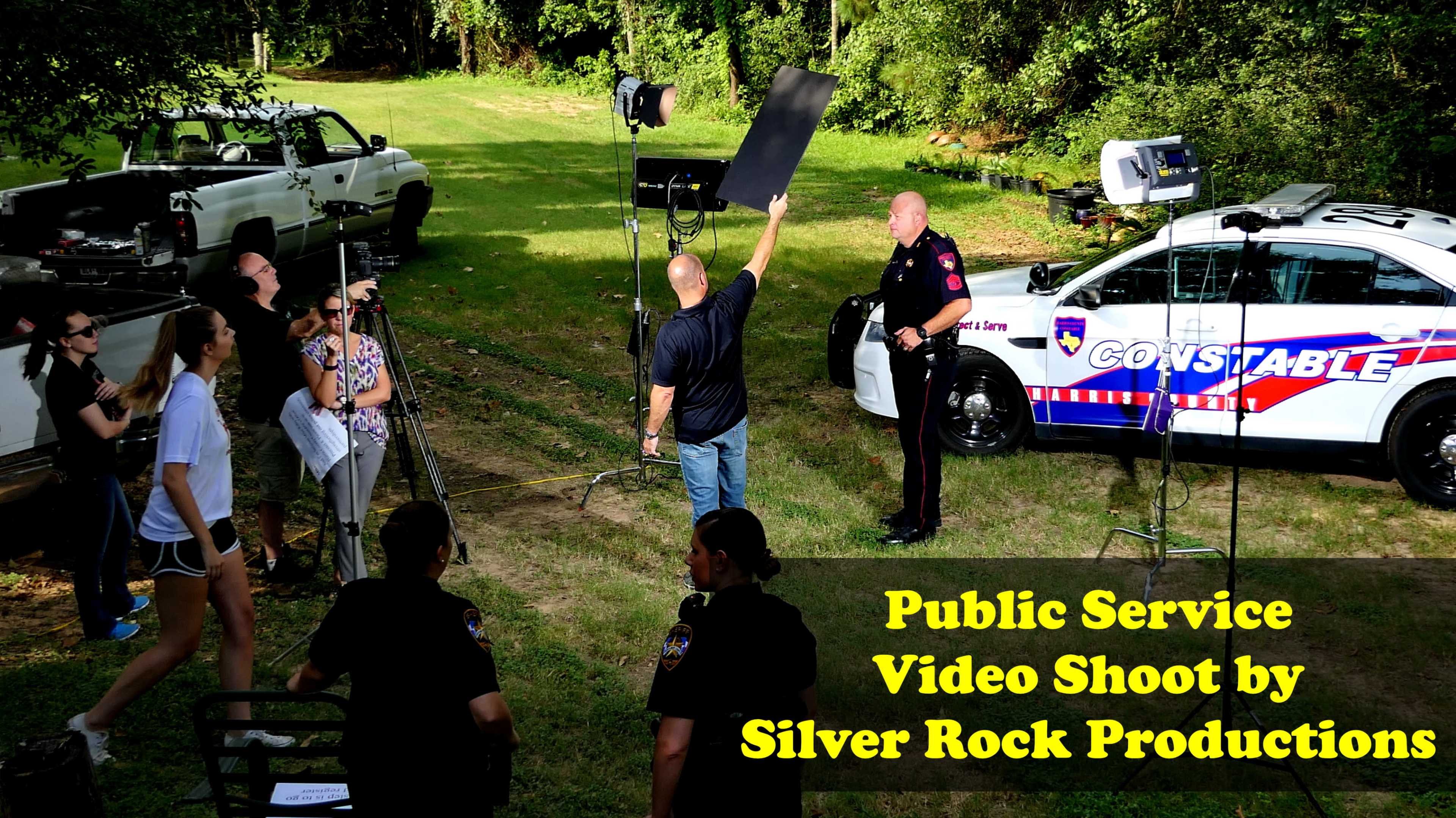 A group of people is setting up a public service video shoot featuring law enforcement personnel beside a marked police vehicle in a wooded area.