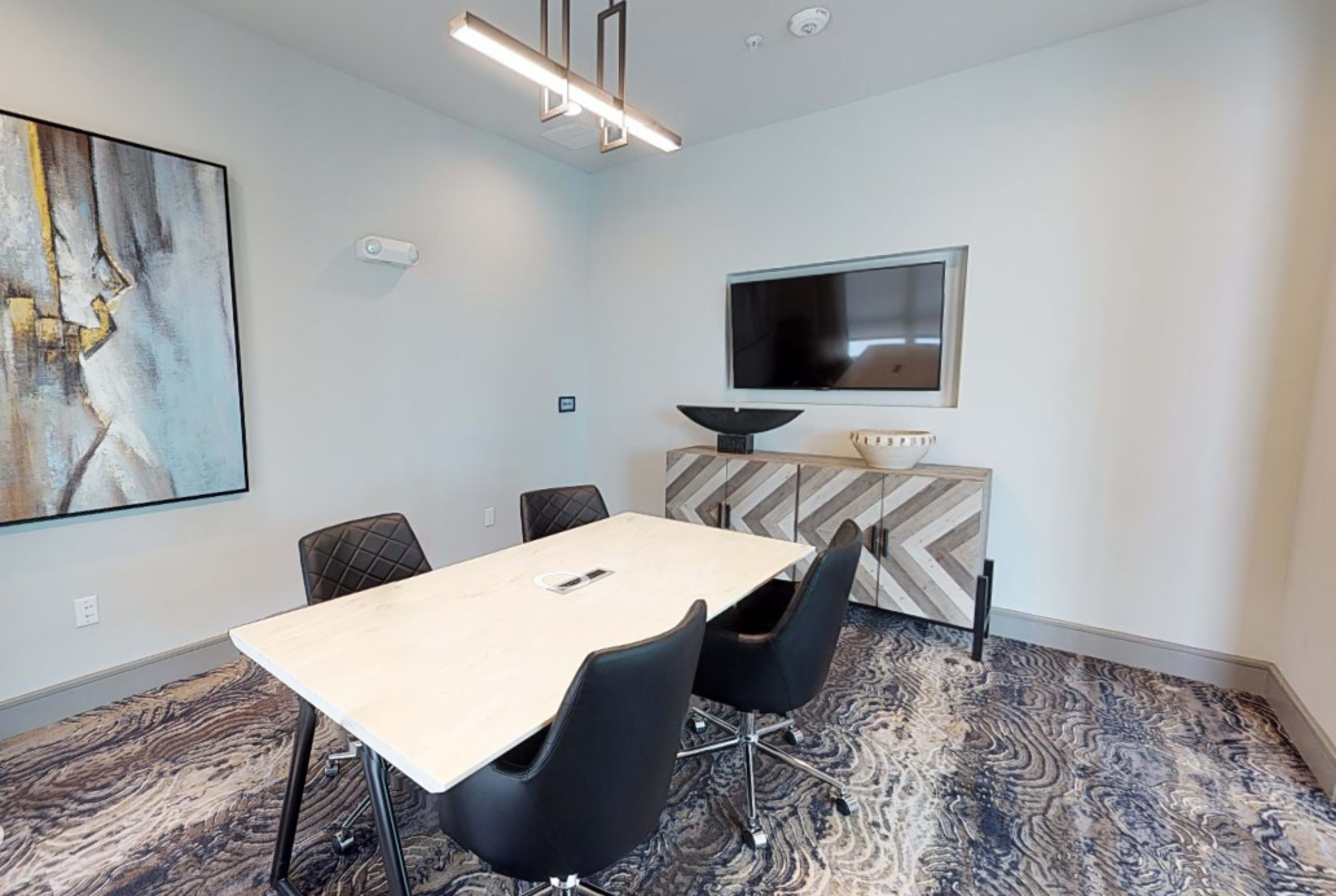 A modern conference room with a rectangular table, four black chairs, a wall-mounted TV, and decorative artwork.