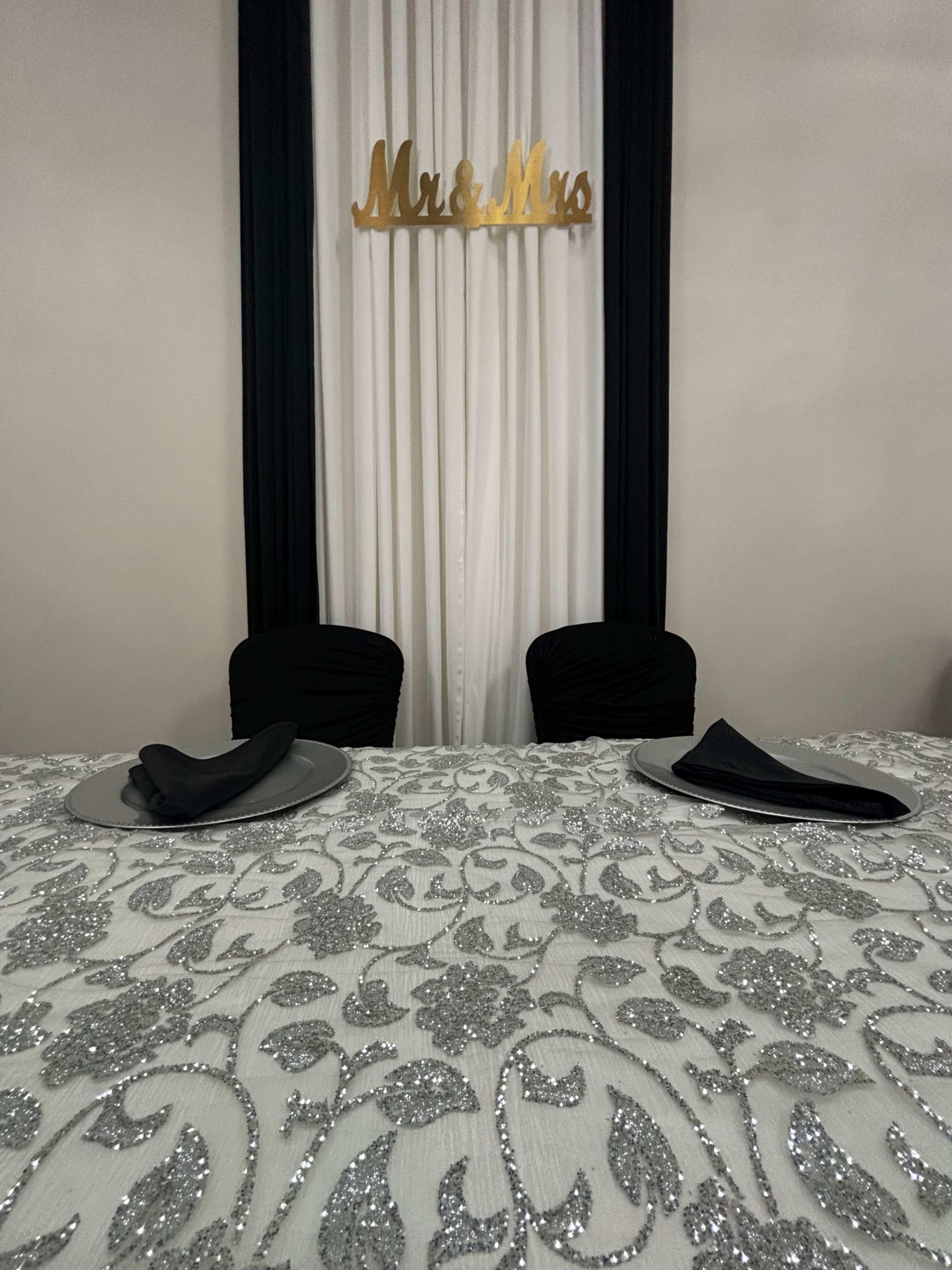 A decorated table with a sparkling tablecloth, two black chairs, and a "Mr. & Mrs." sign hanging above the backdrop.