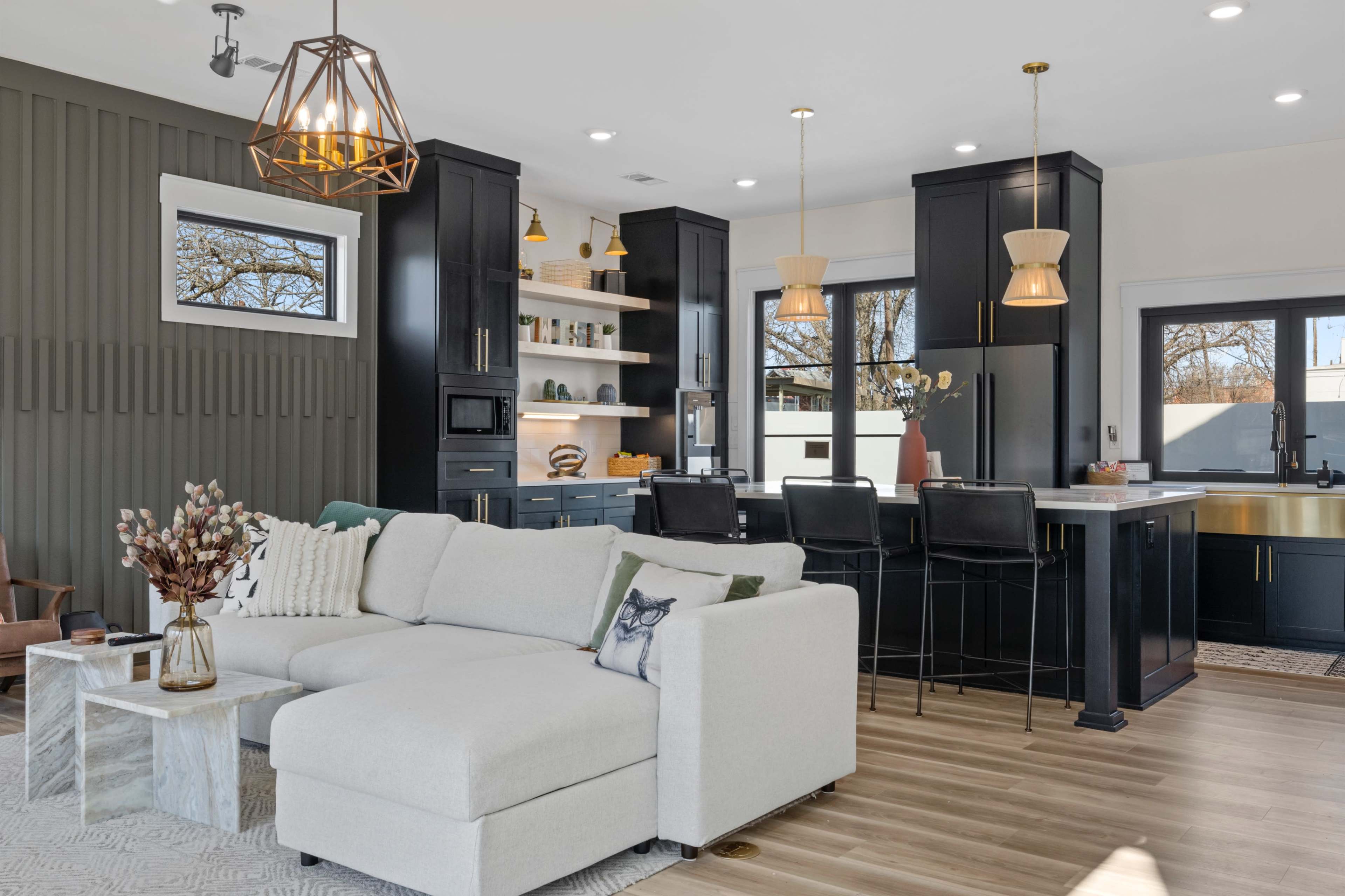 A modern living space features a light-colored sectional sofa, a minimalist coffee table, and a kitchen with dark cabinetry and pendant lighting.