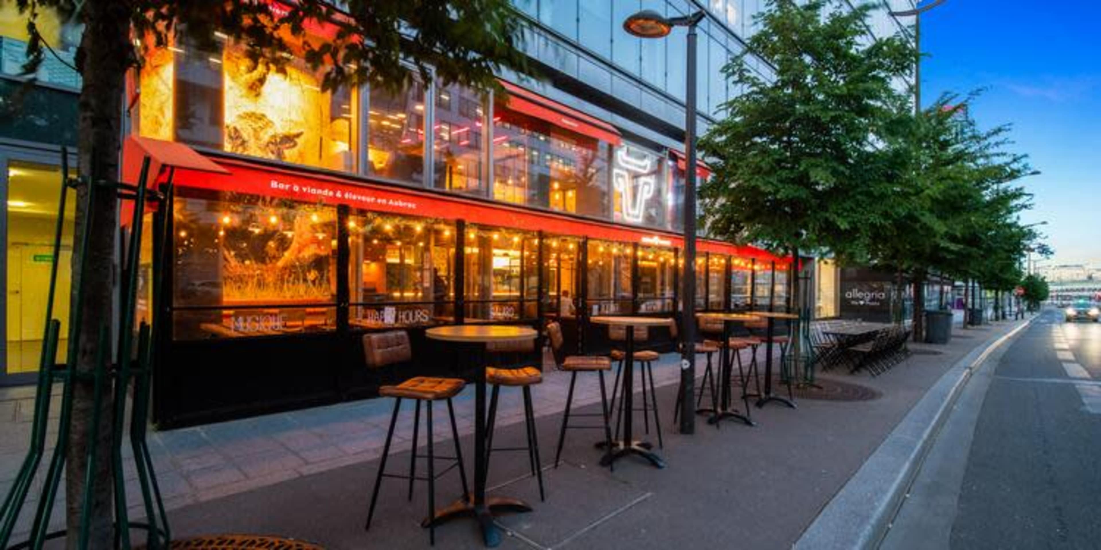 An outdoor seating area of a restaurant with high tables and chairs along a walkway, featuring large windows and a red awning.
