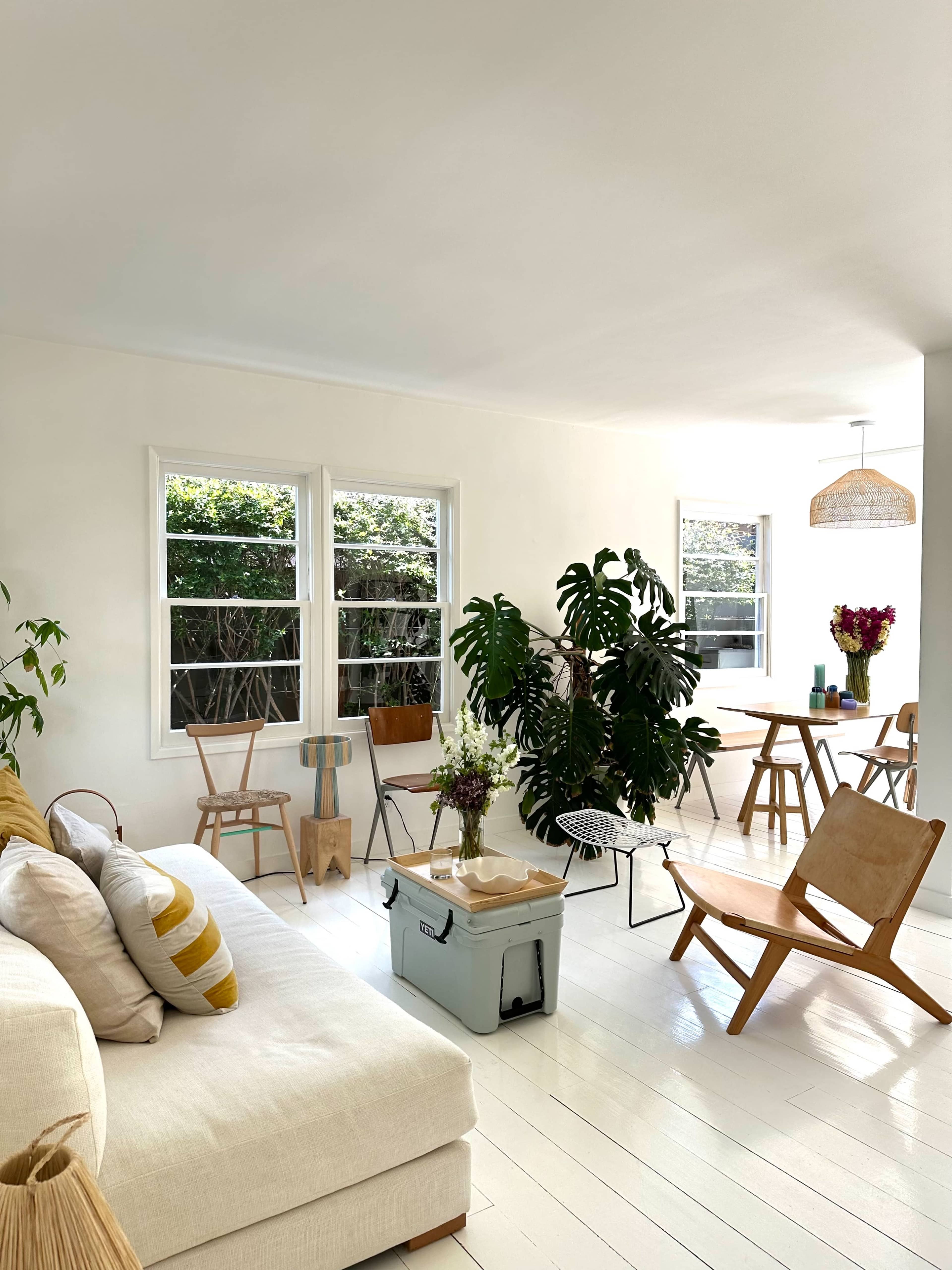 The image depicts a bright, modern living area with white walls, a light sofa, plants, and a dining table surrounded by chairs.