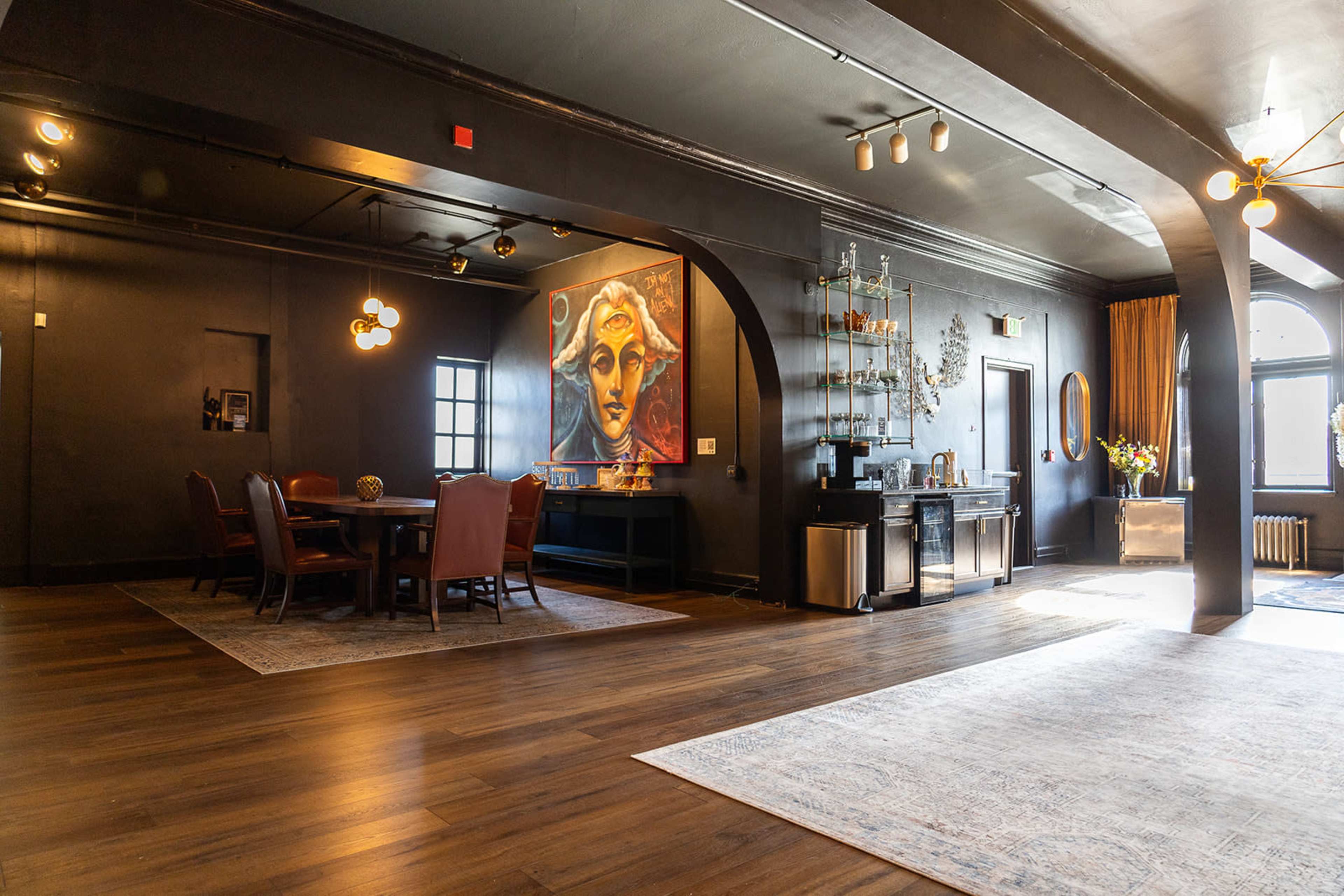 The image shows a spacious room with dark walls, a large mural of a face, a dining area with a wooden table and chairs, and a sideboard with decorative items.