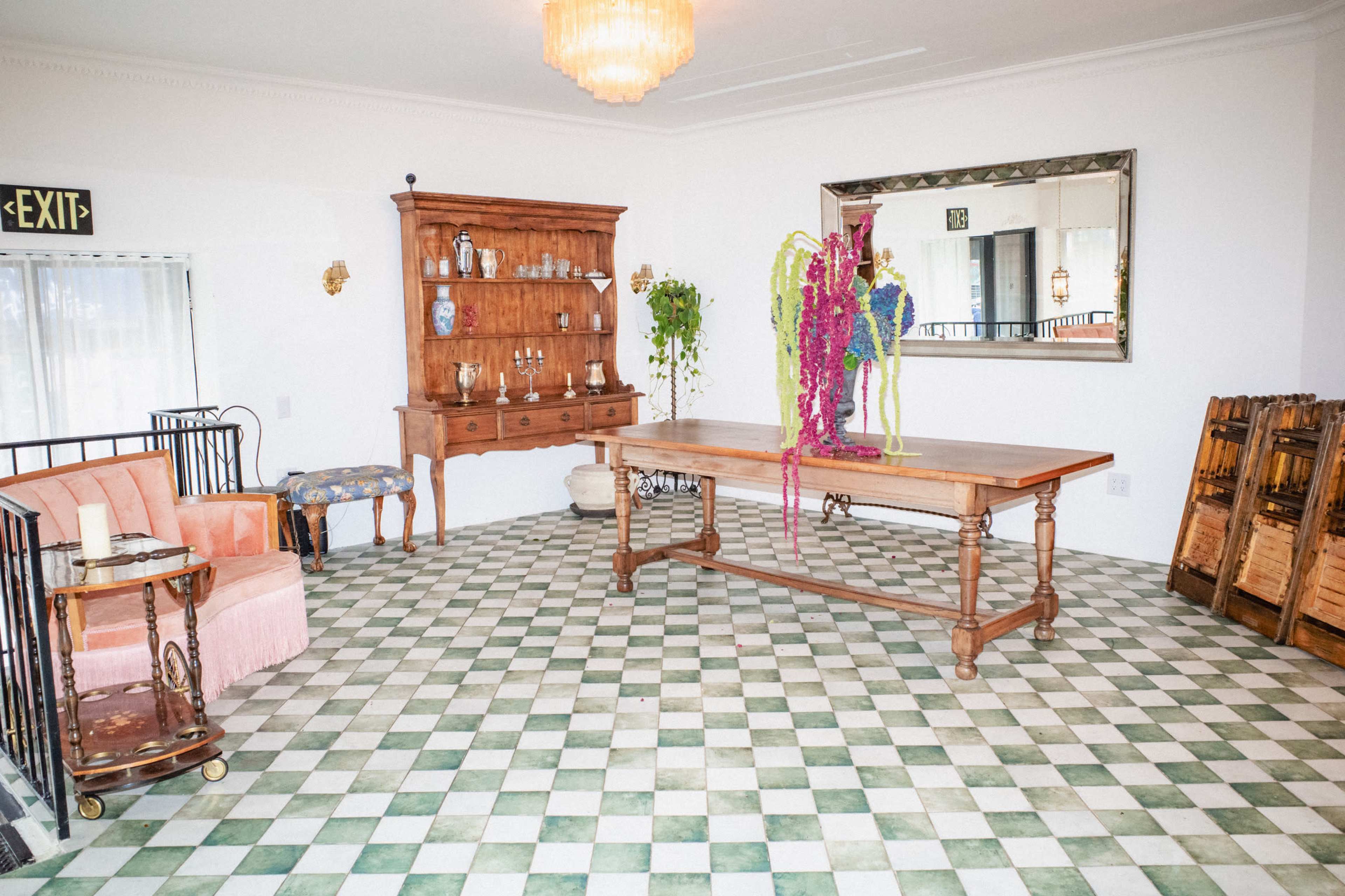 The room features a wooden table at its center, surrounded by patterned flooring, with a vintage cabinet and decorative flowers displayed prominently.