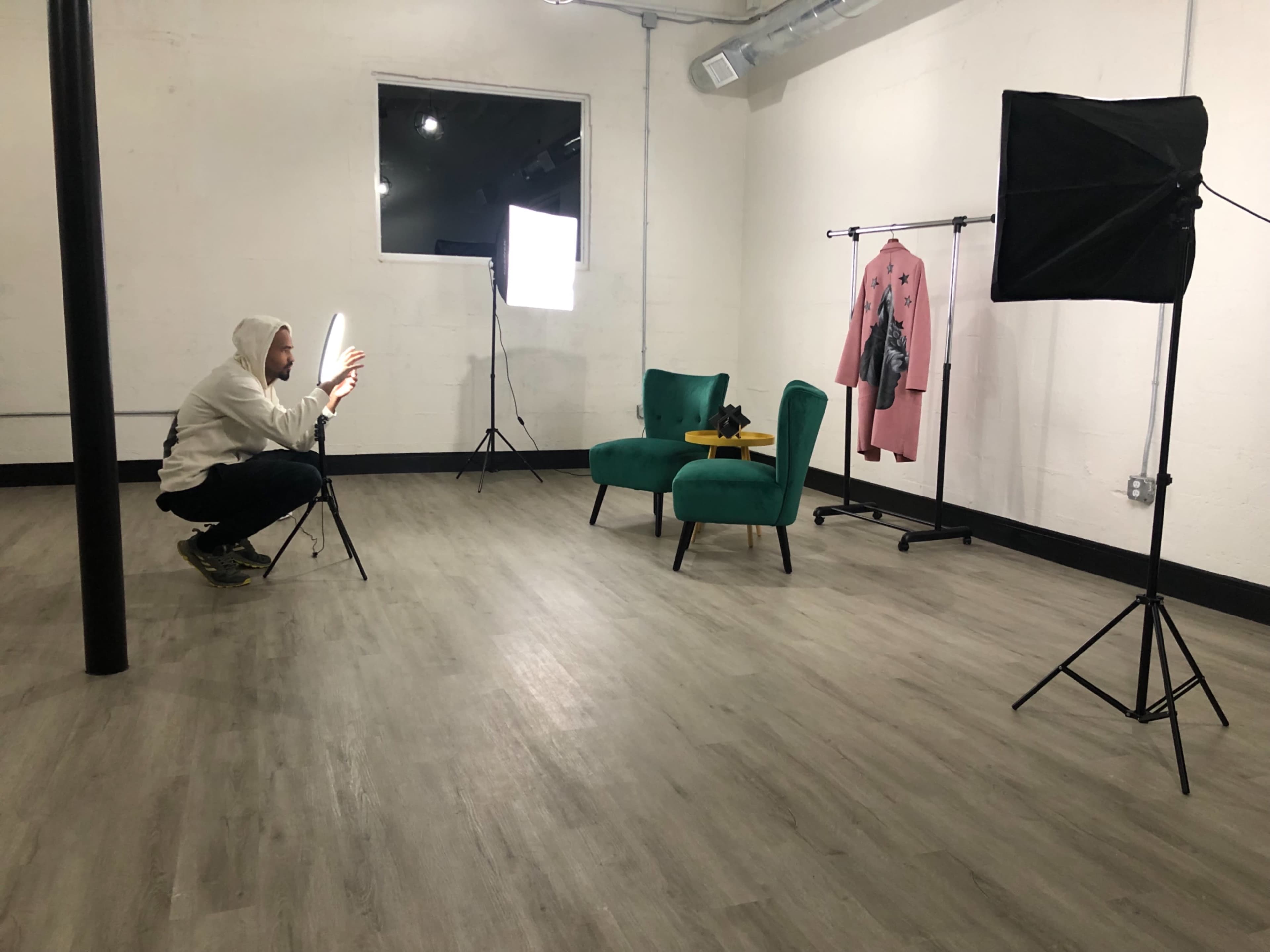 A person in a hoodie is crouched next to a chair in a photography studio, adjusting lighting equipment while a rack with clothing stands nearby.