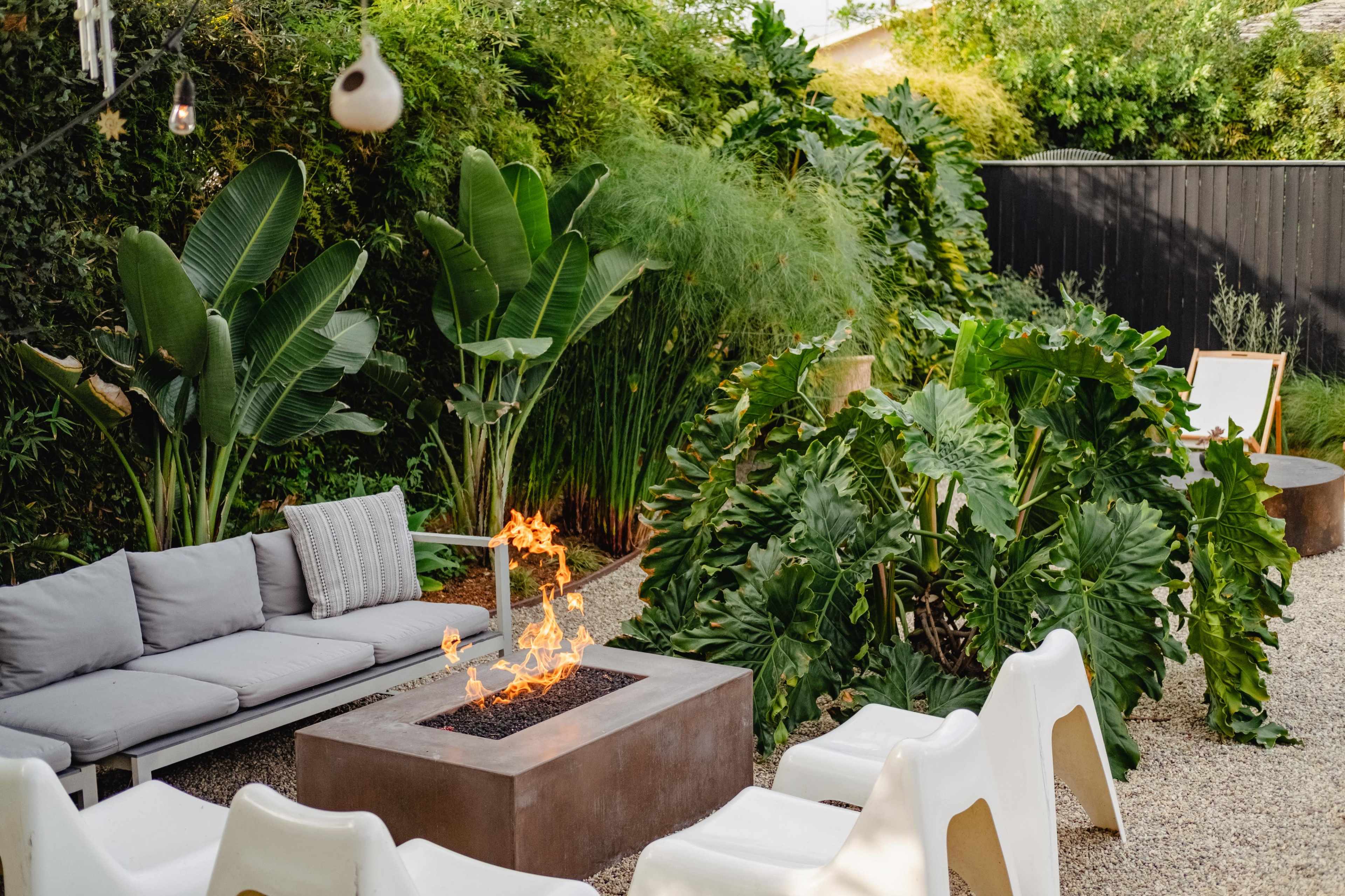 A modern outdoor seating area features a fire pit surrounded by white chairs and lush green plants.