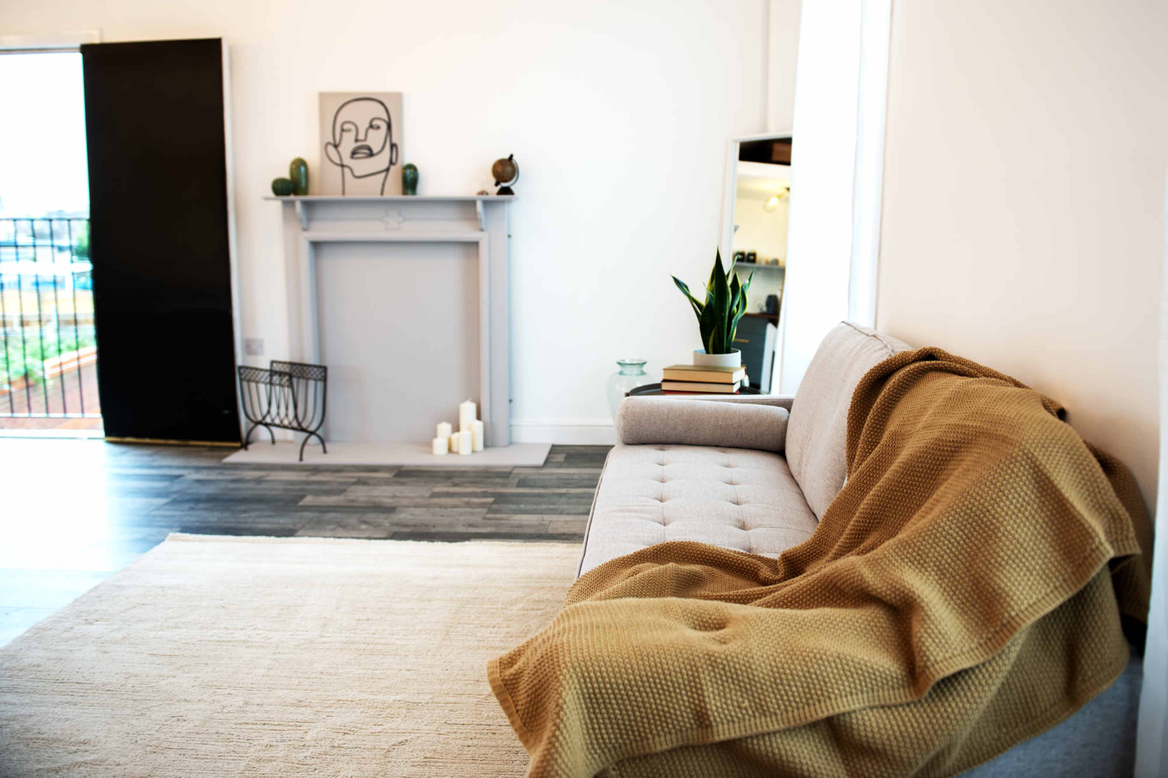 A modern living room with a gray couch draped with a brown blanket, a minimalist fireplace, a plant, and a mirror reflecting the interior.