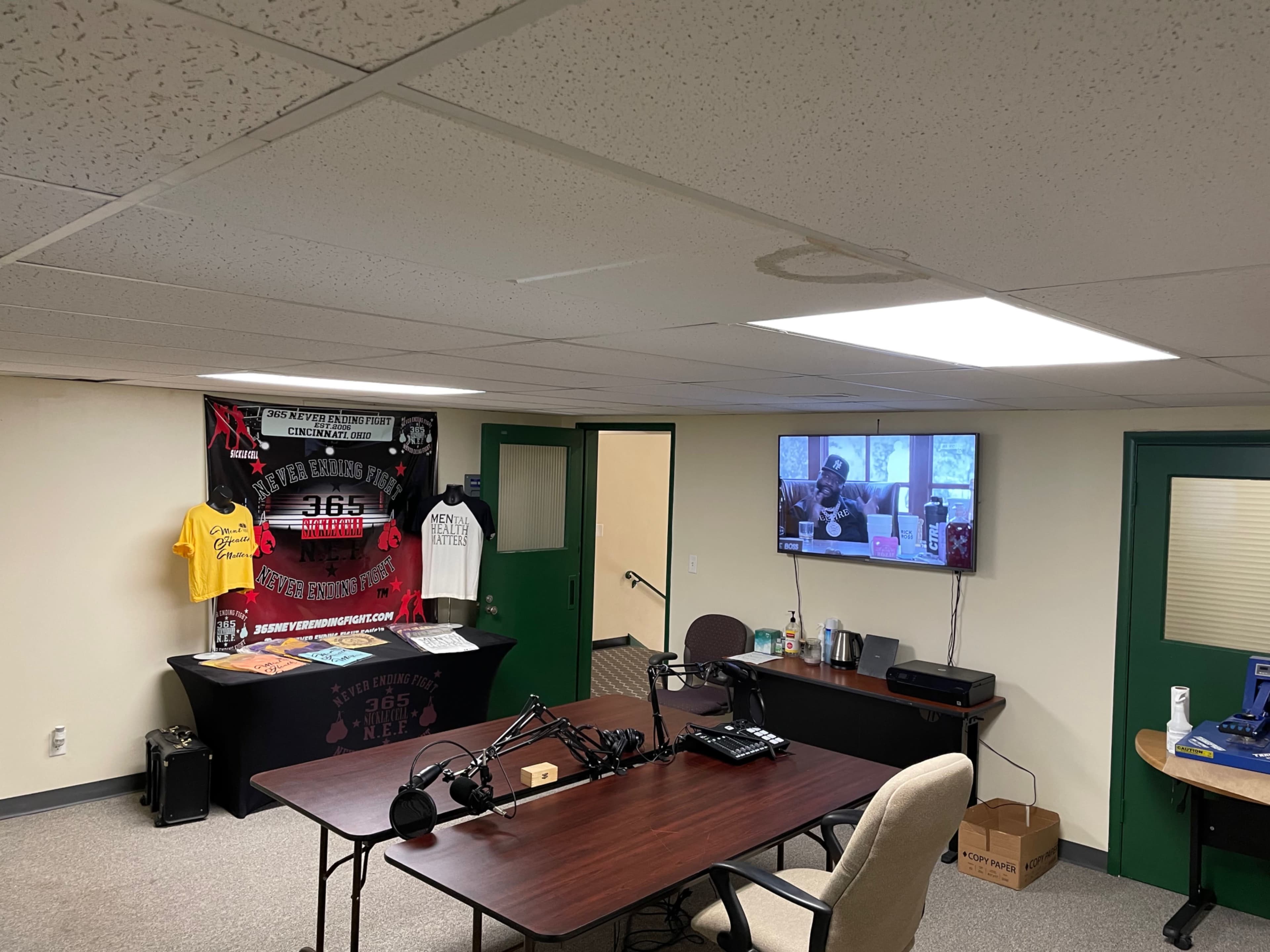 The image shows a room with a table and chairs arranged for a podcast setup, featuring a microphone, a television displaying a video, and a wall adorned with promotional materials and clothing.