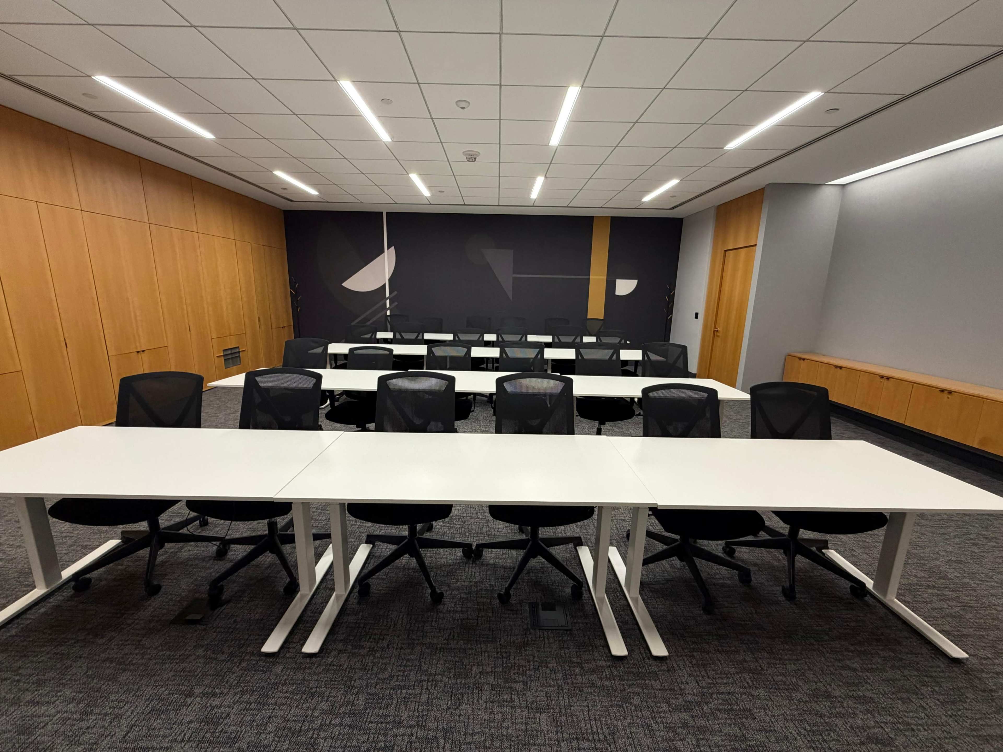 The image shows a modern conference room with several long white tables arranged in rows and black ergonomic chairs.