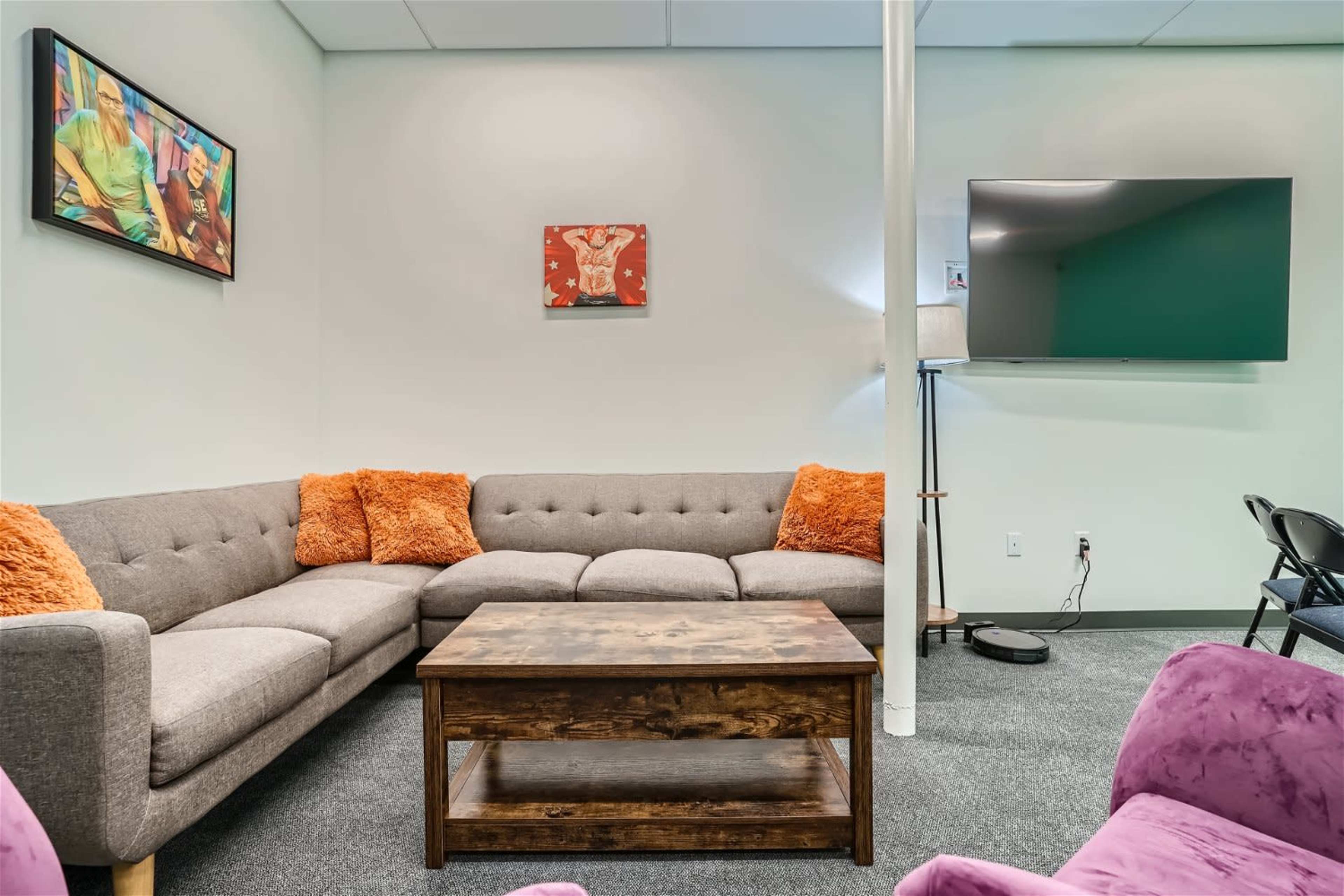 A modern lounge area with a gray sectional sofa featuring orange cushions, a wooden coffee table, and a mounted television on the wall.