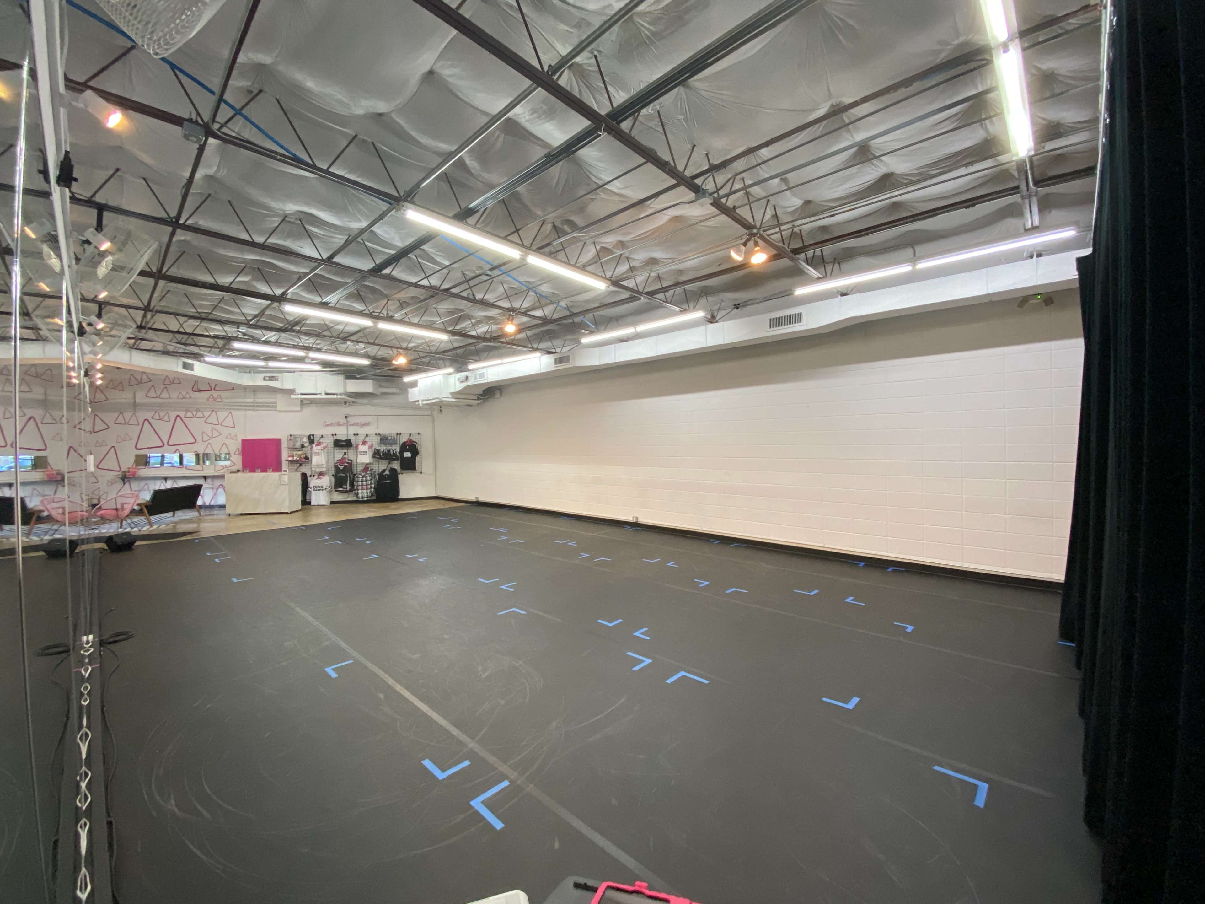 The image shows a spacious dance studio with black flooring marked by blue tape and a wall lined with mirrors and equipment.