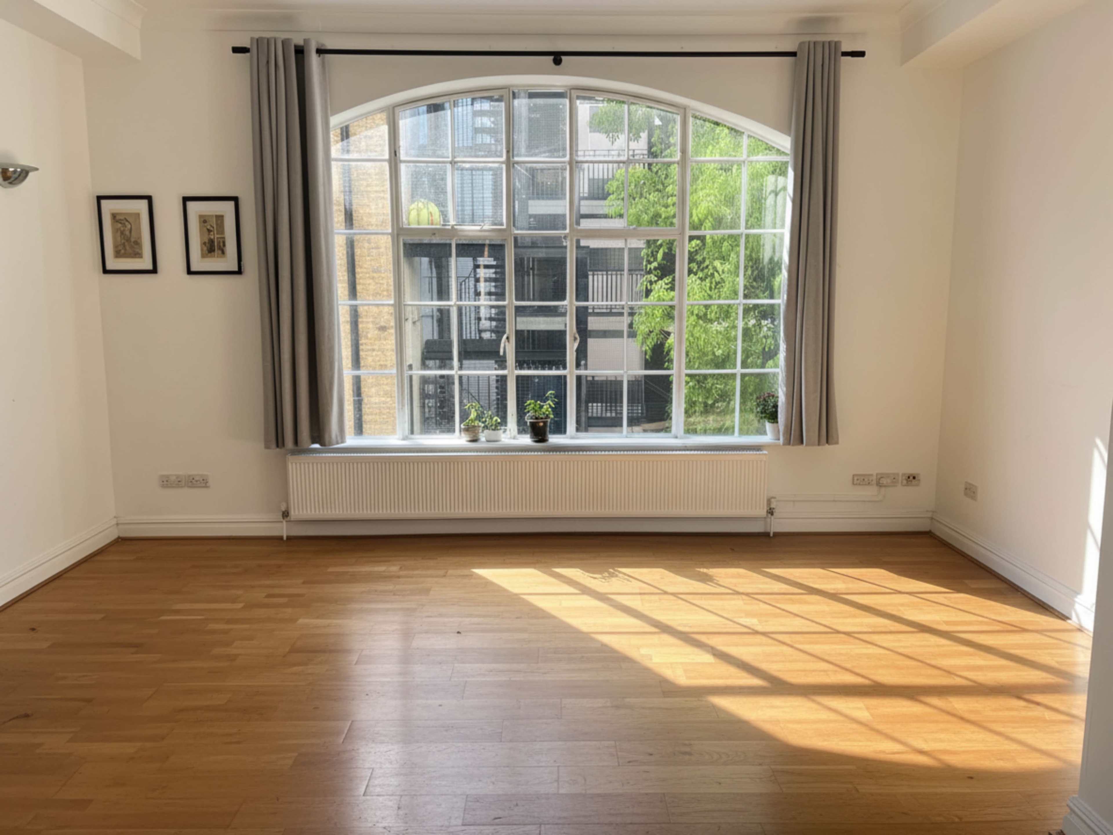A bright, empty room with wooden flooring and large arched windows overlooking a green view.