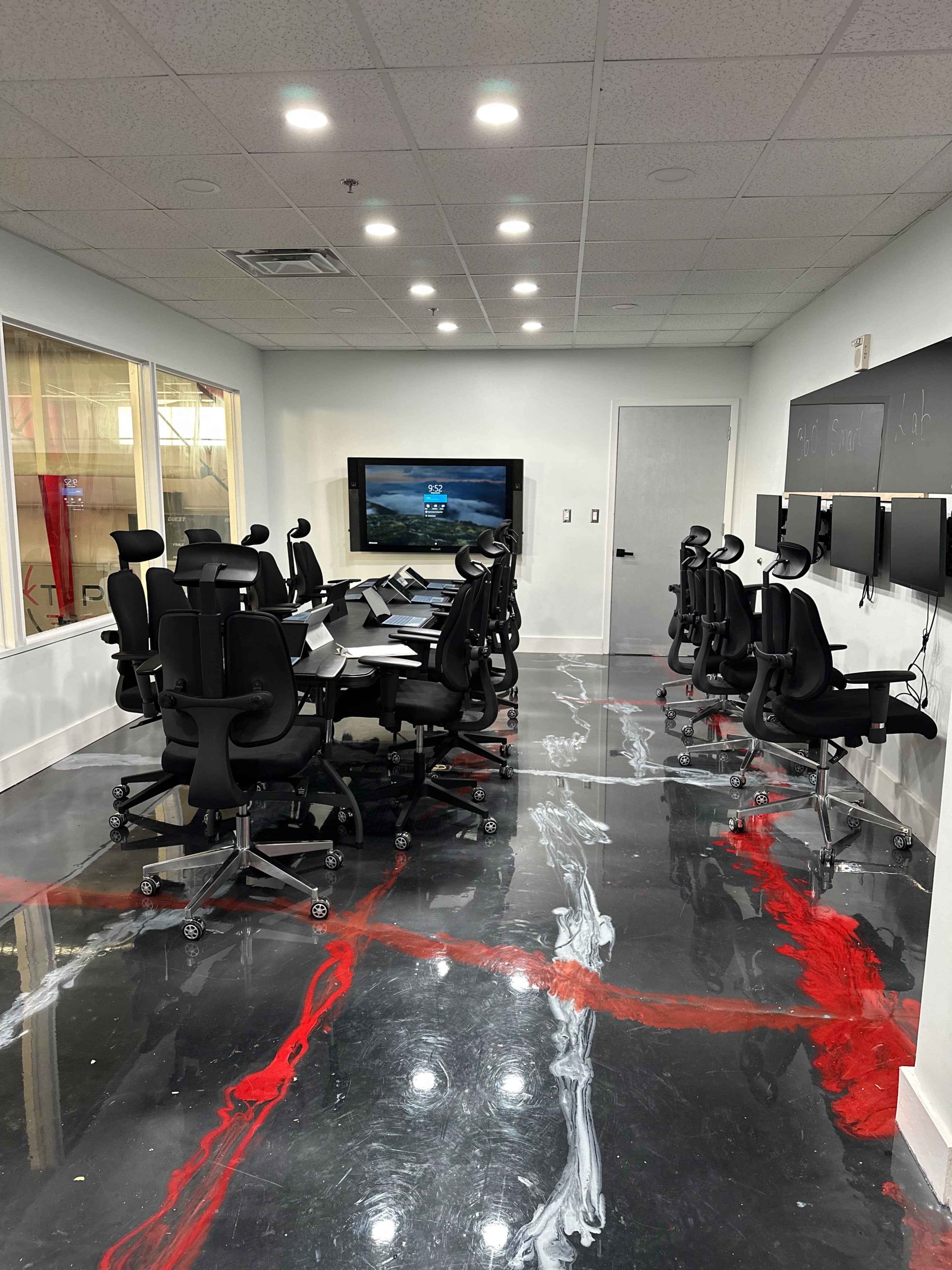 The image shows a modern conference room with several ergonomic chairs arranged around a long table, a large screen mounted on the wall, and a chalkboard.