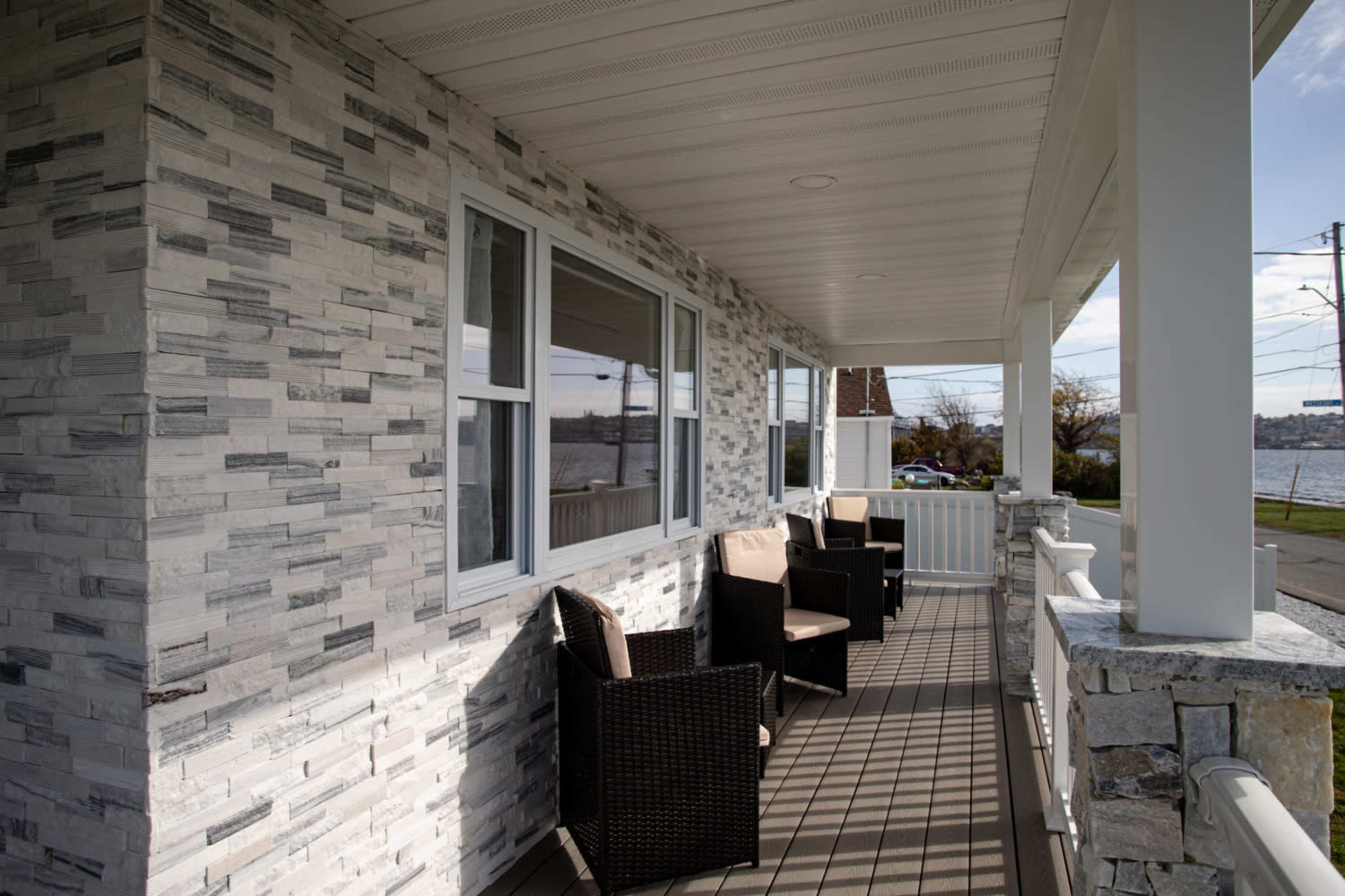 A covered porch with woven chairs and a stone-clad wall overlooking a body of water.