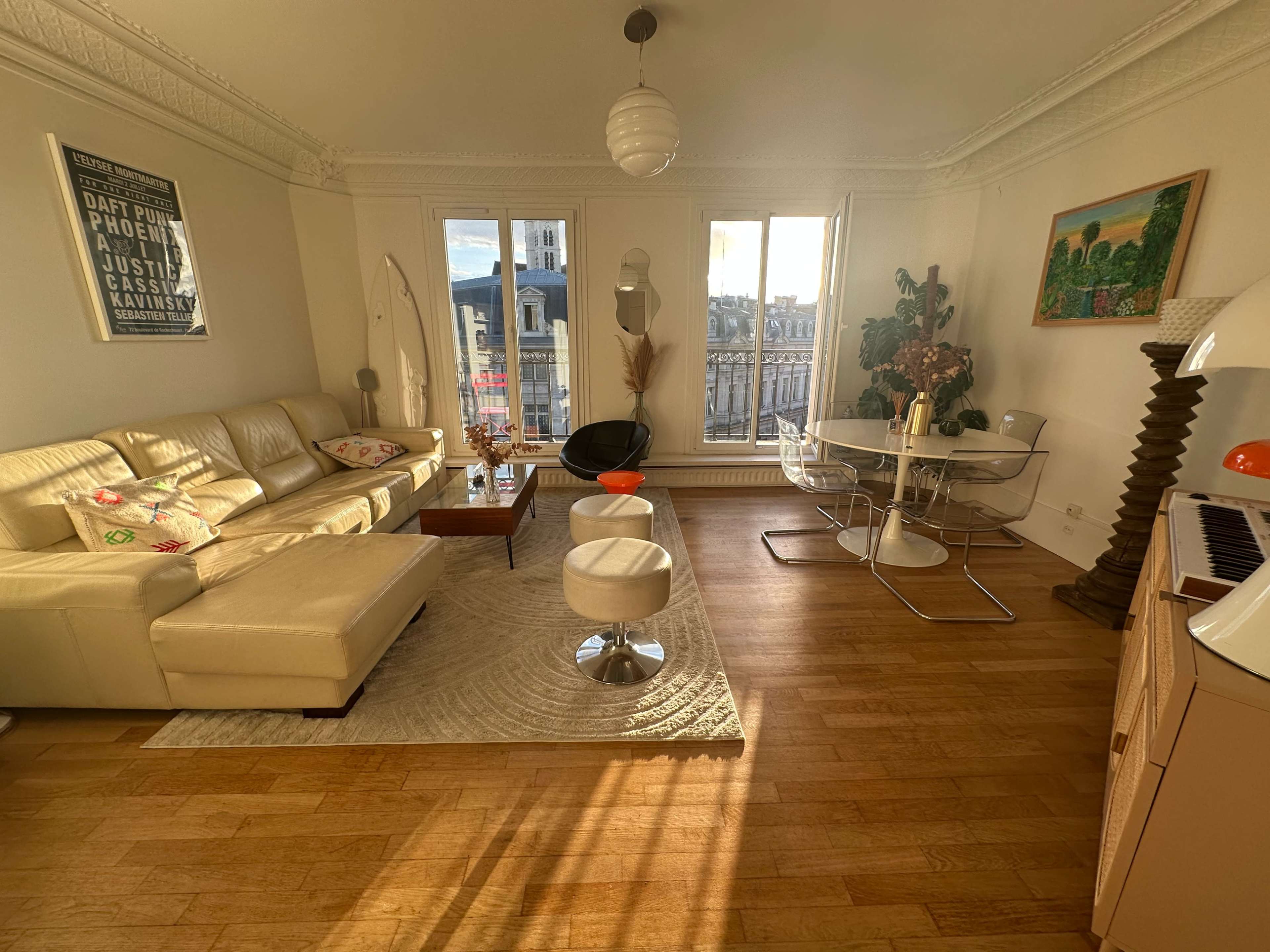A modern living room features a cream leather sofa, a glass dining table, and large windows that let in natural light, with decorative elements on the walls and floor.