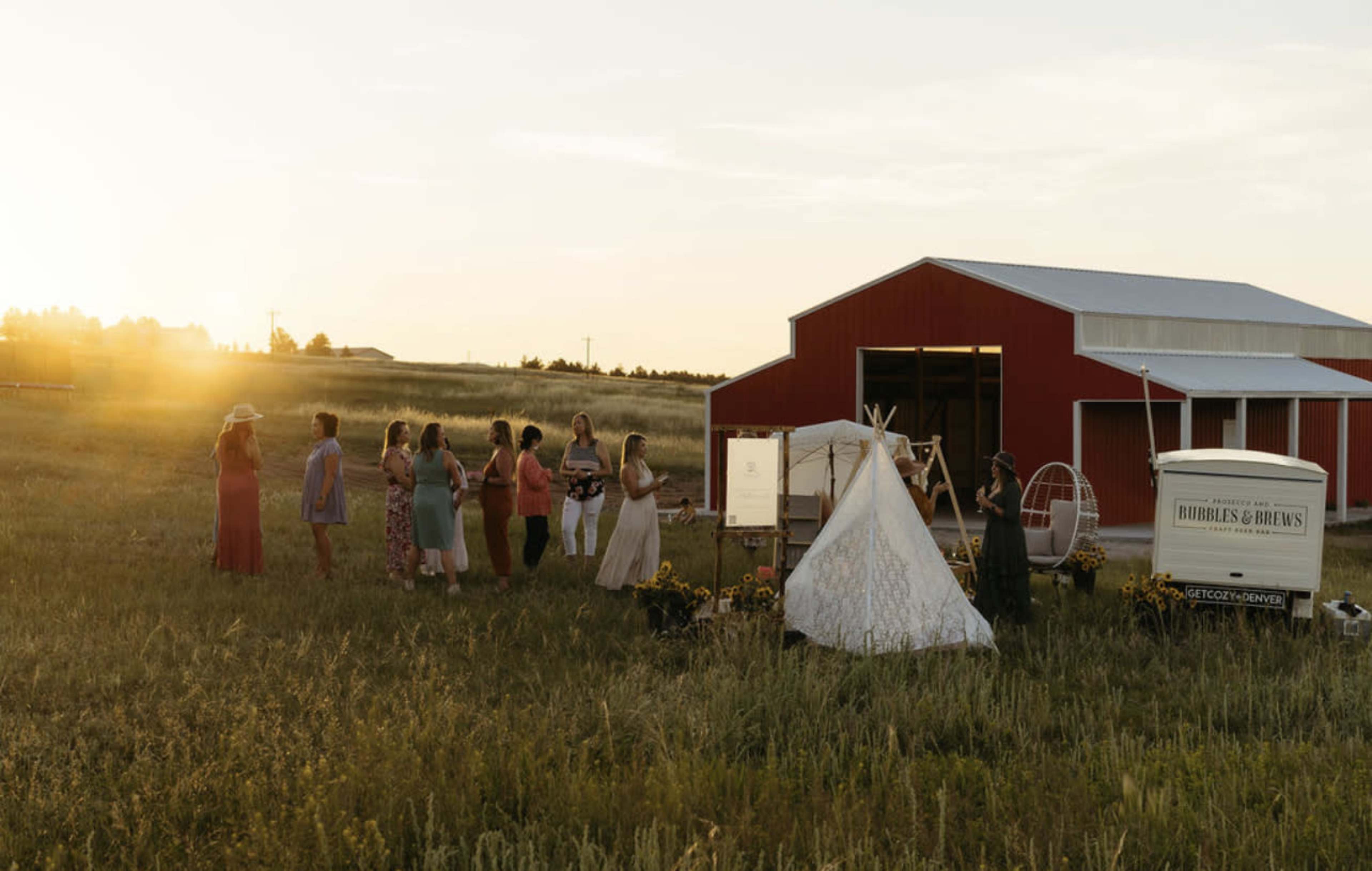 A group of women gathers in a field near a red barn during sunset, with a white tent set up nearby.