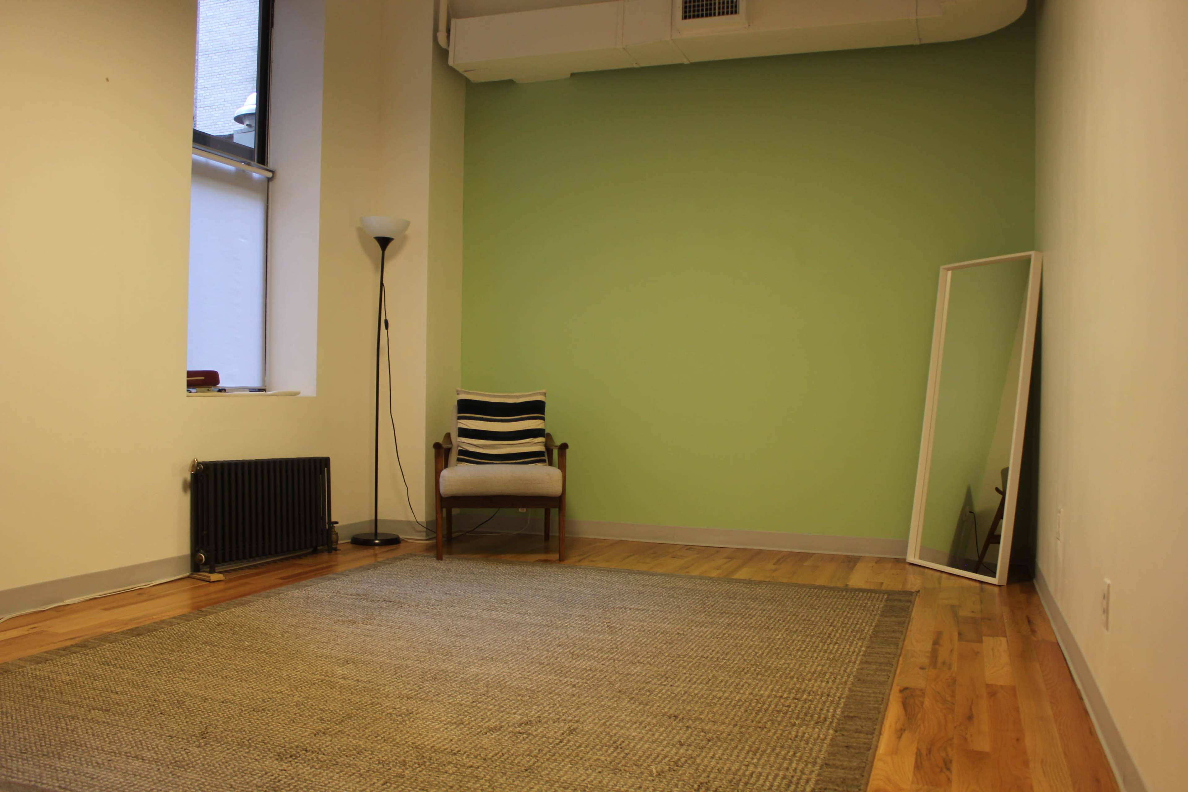 A minimalist room with a green accent wall, a chair with a striped cushion, a floor lamp, a large mirror, and wooden flooring.