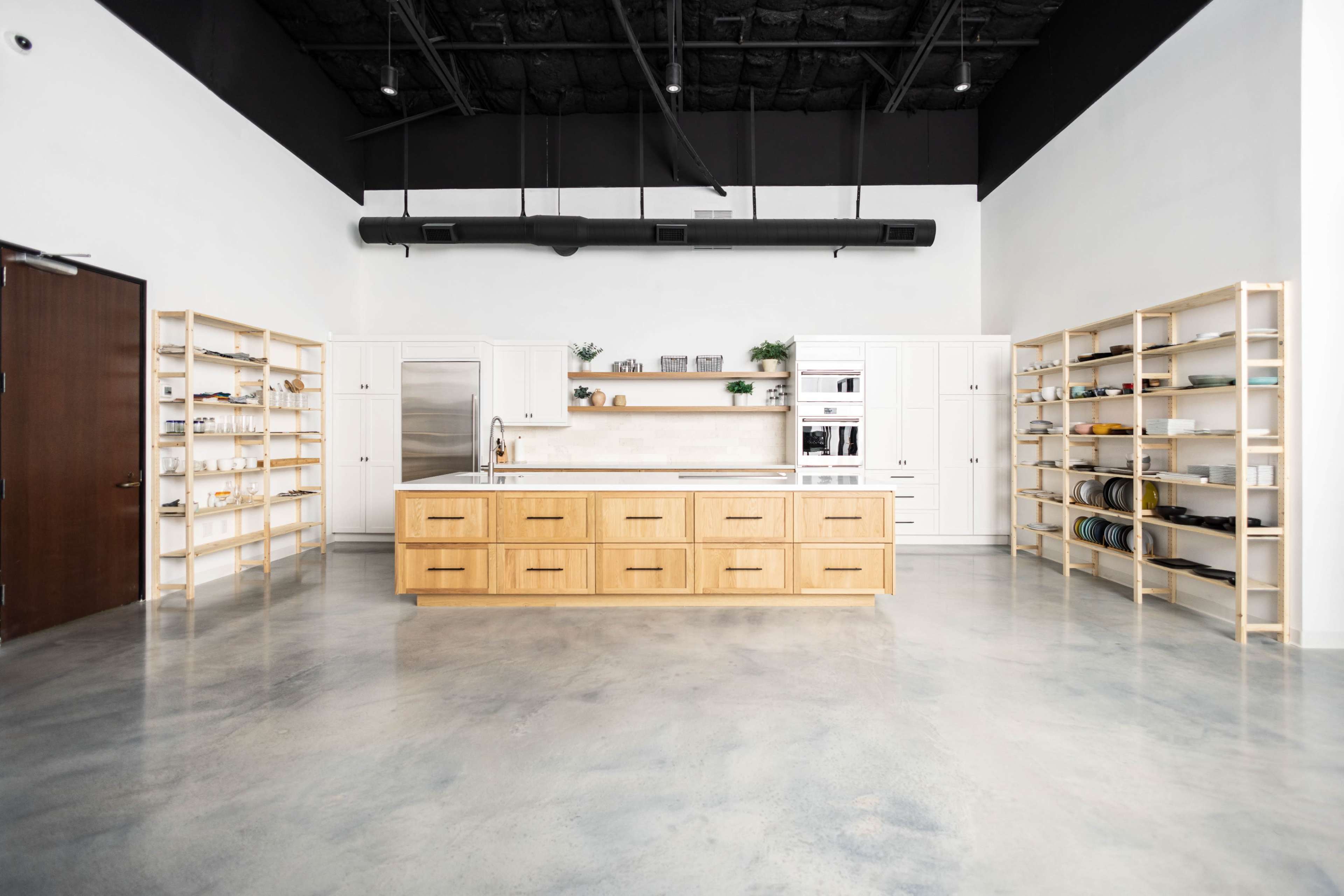 A spacious modern kitchen features a central wooden island with storage drawers, surrounded by open shelving and white cabinetry against a stark black ceiling.