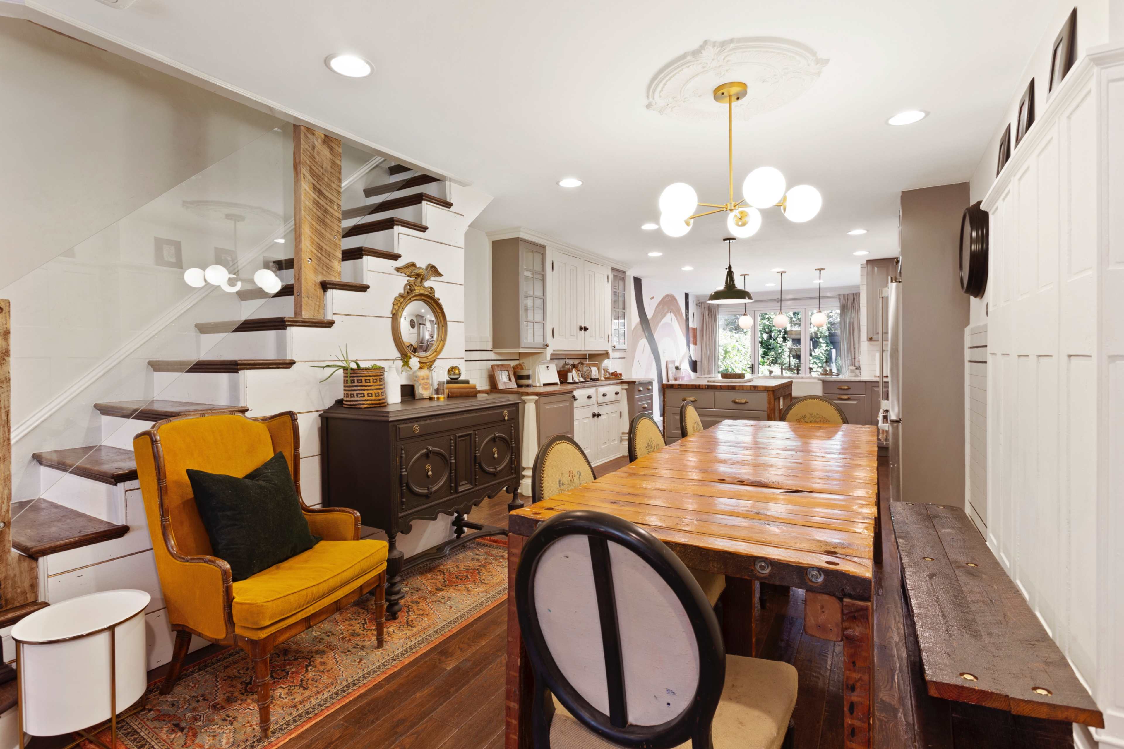 A modern kitchen and dining area featuring a wooden dining table, an ornate light fixture, and a staircase with a glass railing.