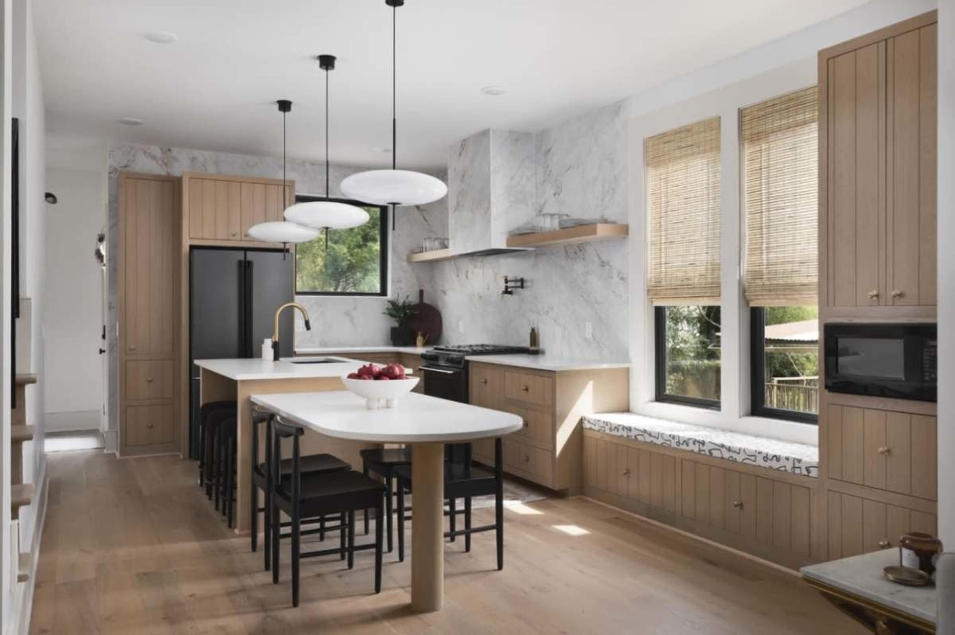 A modern kitchen features light wood cabinetry, a large island with seating, and large windows allowing natural light to fill the space.