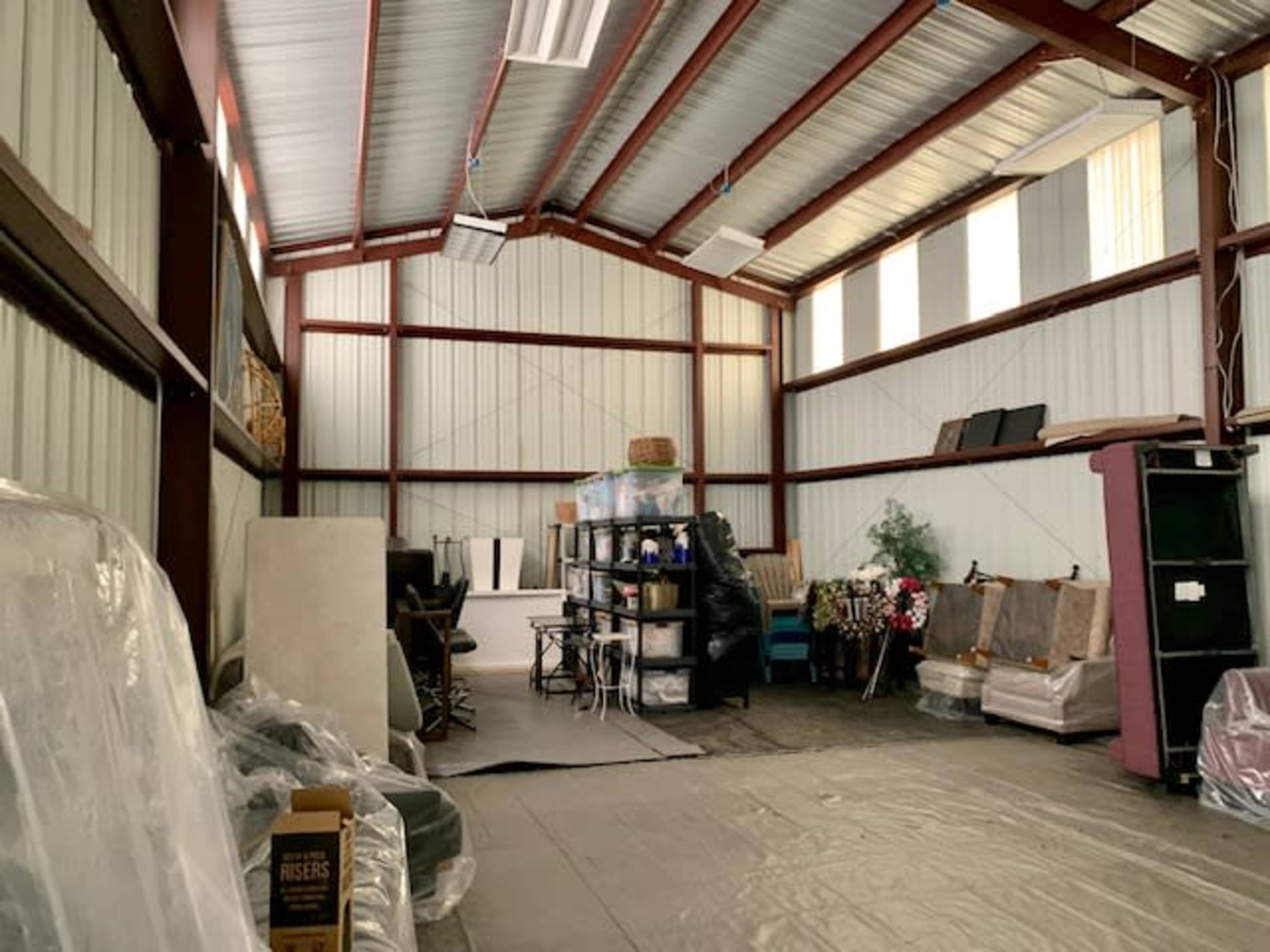 An industrial storage space with metal walls and a high ceiling, filled with various items and furniture stacked along the sides.