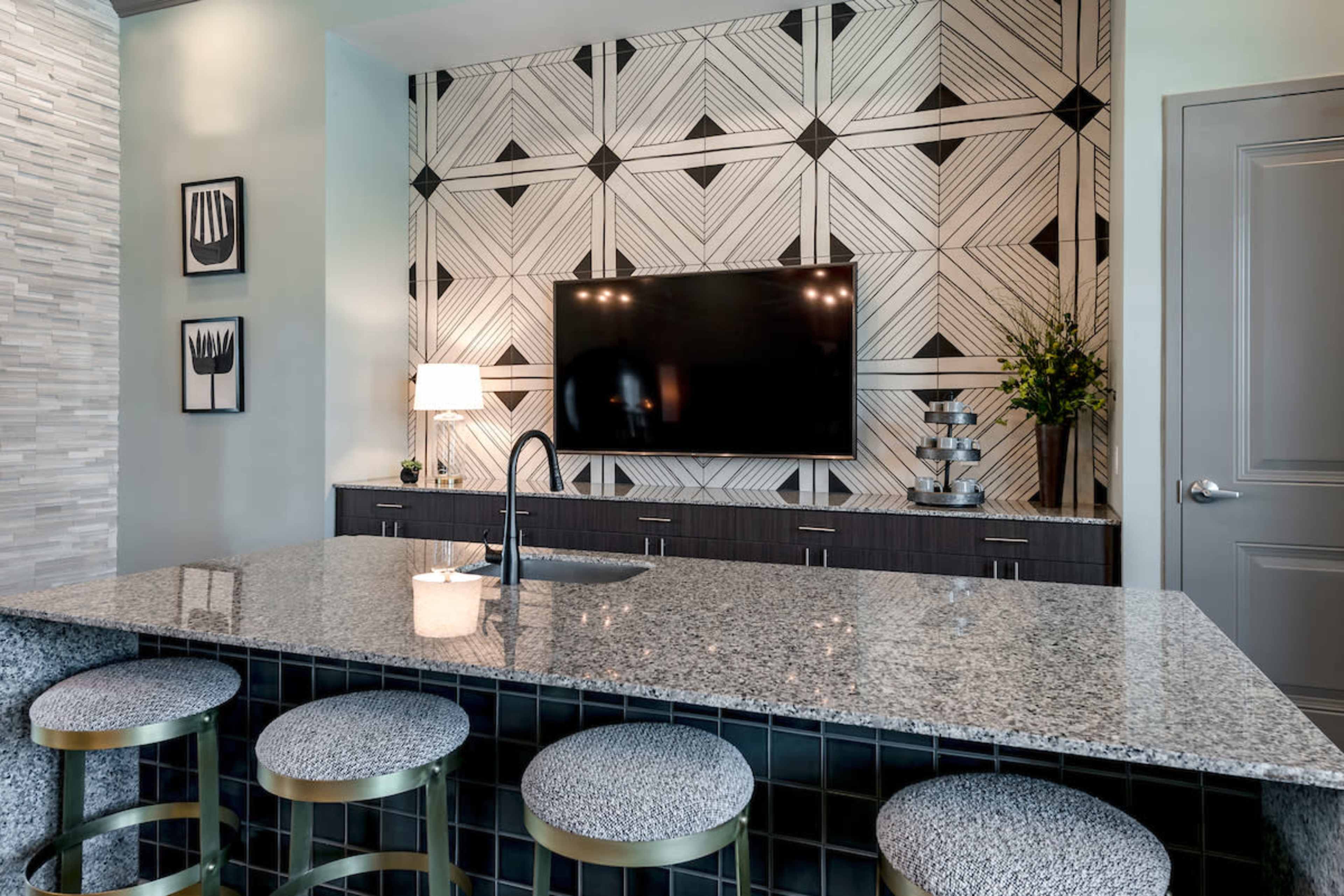 A modern kitchen features a granite island with four upholstered stools, a large wall-mounted TV, and a geometric patterned accent wall behind dark cabinetry.
