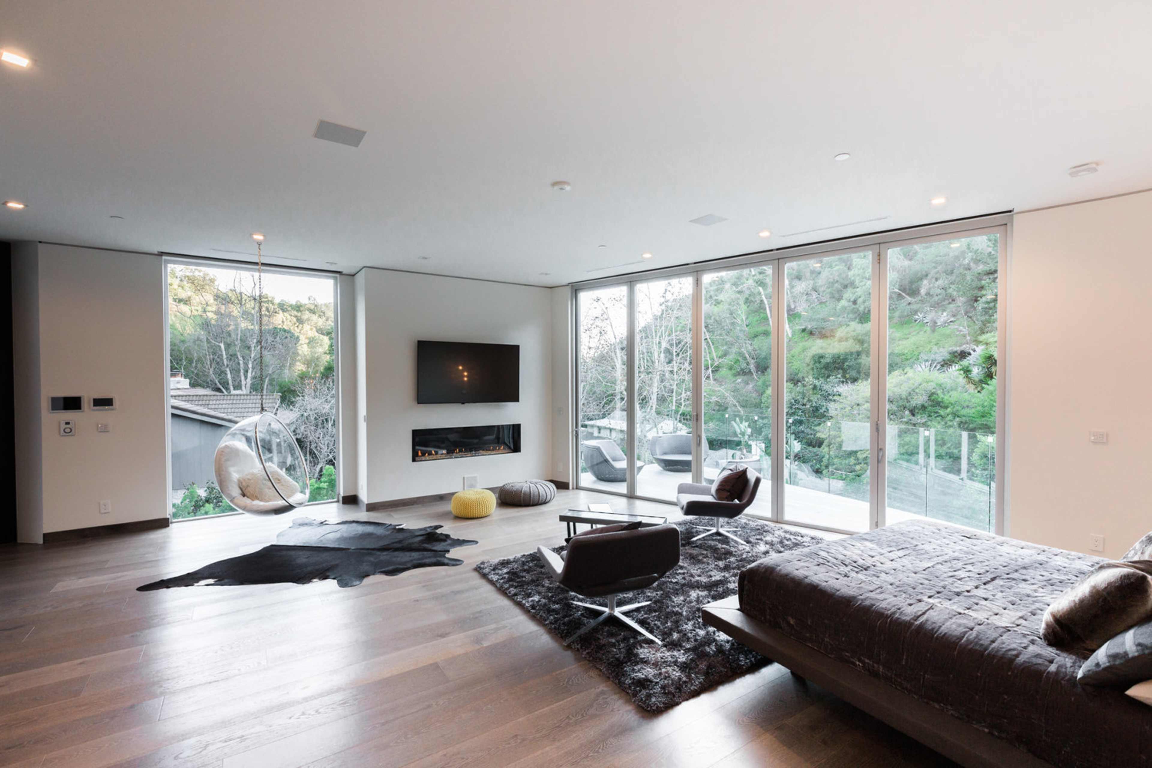 A modern living room with large windows overlooking a green landscape, featuring a hanging chair, a flat-screen TV, and sleek furniture on hardwood floors.