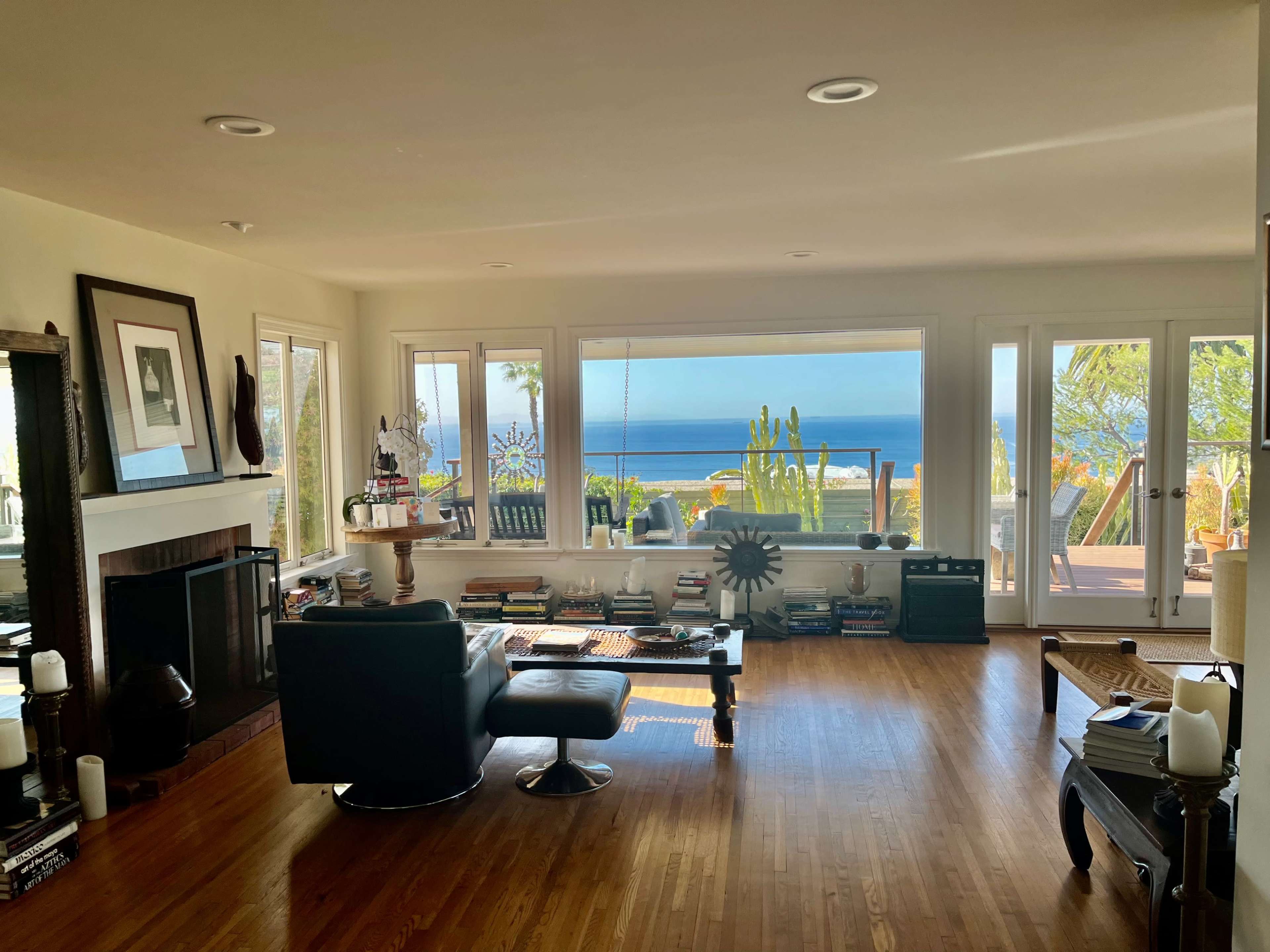 The living room features a large window overlooking the ocean, with a black leather chair, a coffee table stacked with books, and a fireplace.