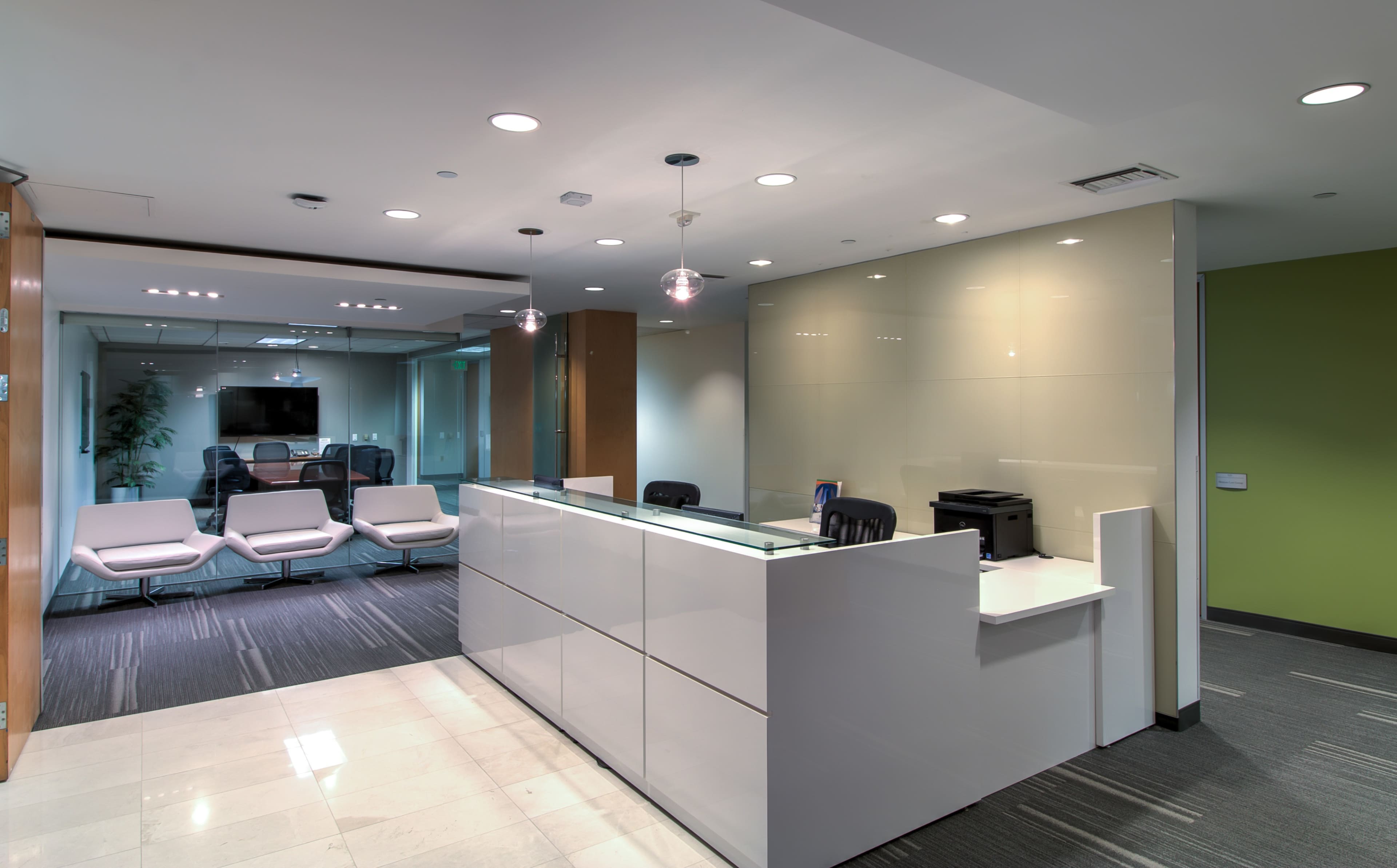 The image shows a modern office reception area with a white reception desk, minimalist seating, and a conference room visible in the background.