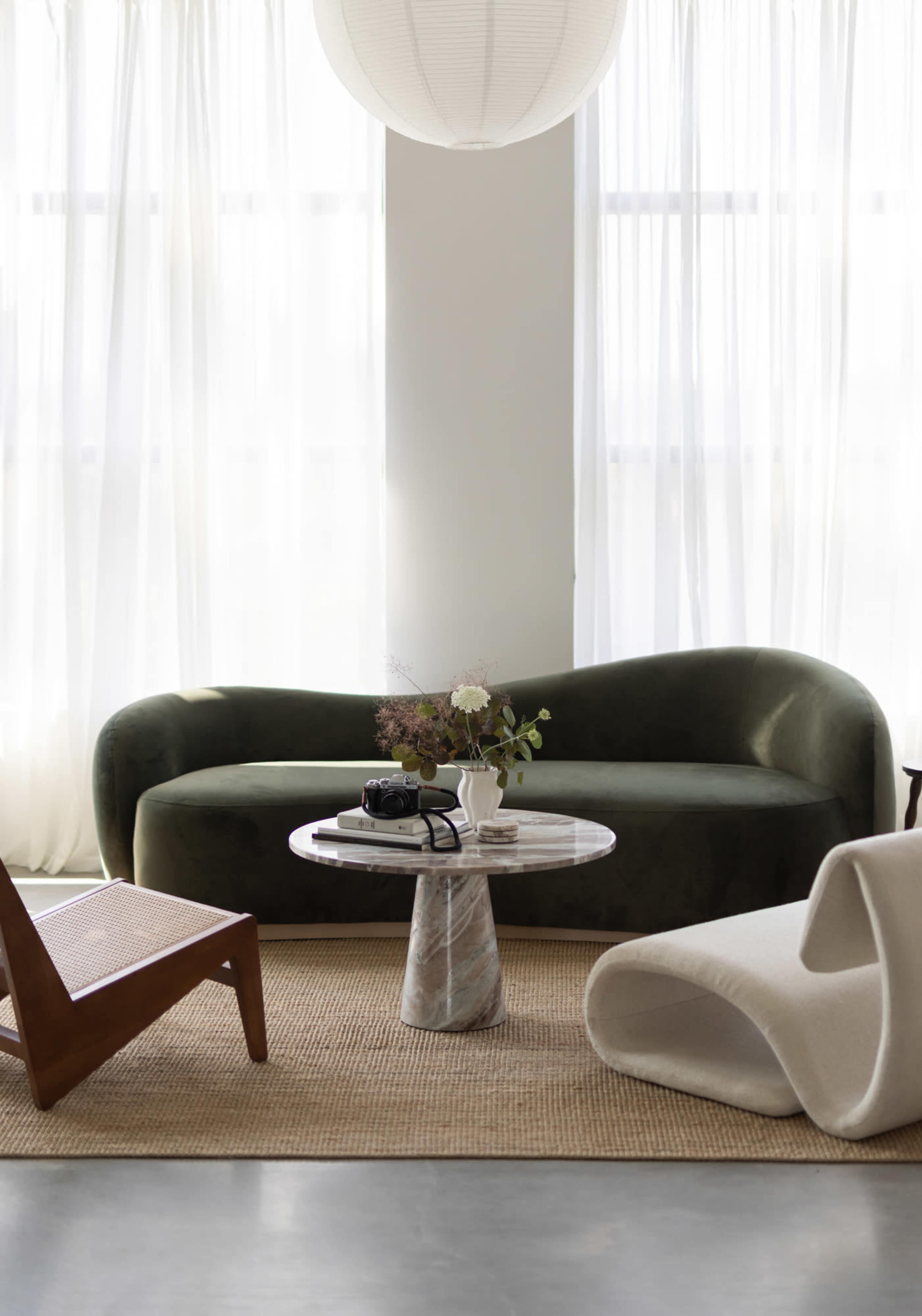 The image features a minimalist living room with a green curved sofa, a round marble coffee table, and a woven rug under soft natural light from large windows.
