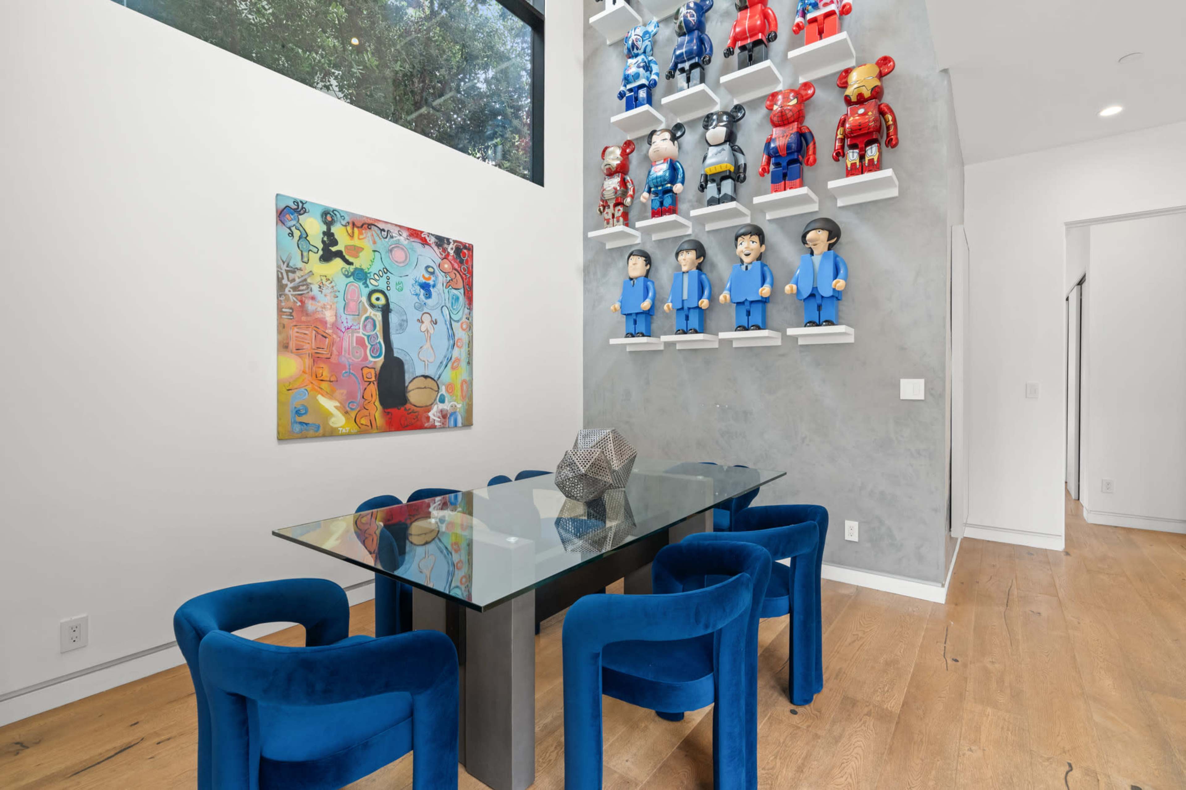 A modern dining area features a glass table surrounded by blue chairs, with a colorful artwork hanging on one wall and a display of figurines on the other.