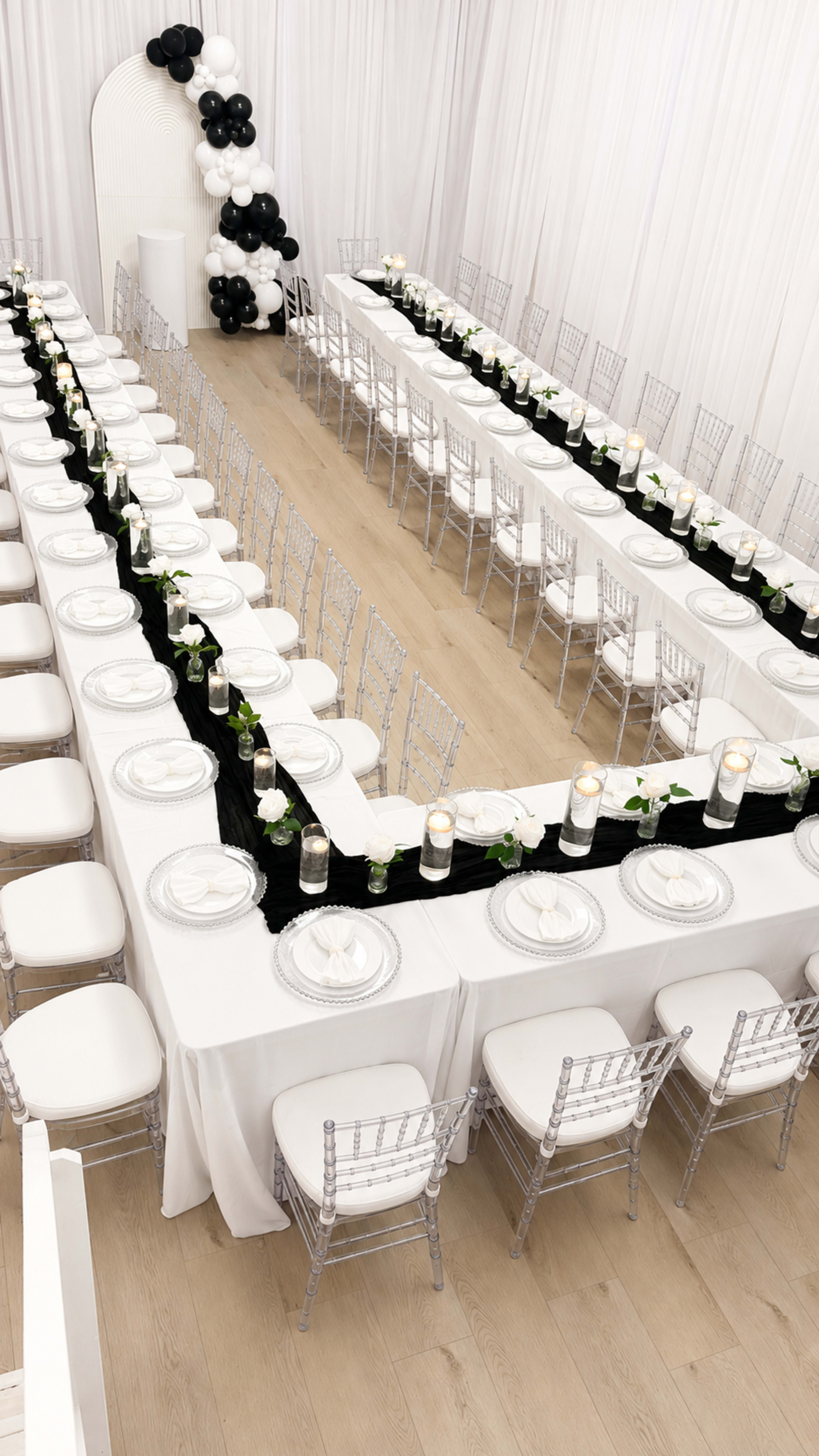The image shows a elegantly arranged banquet hall with long tables set with white plates, black runners, and floral centerpieces, surrounded by transparent chiavari chairs.