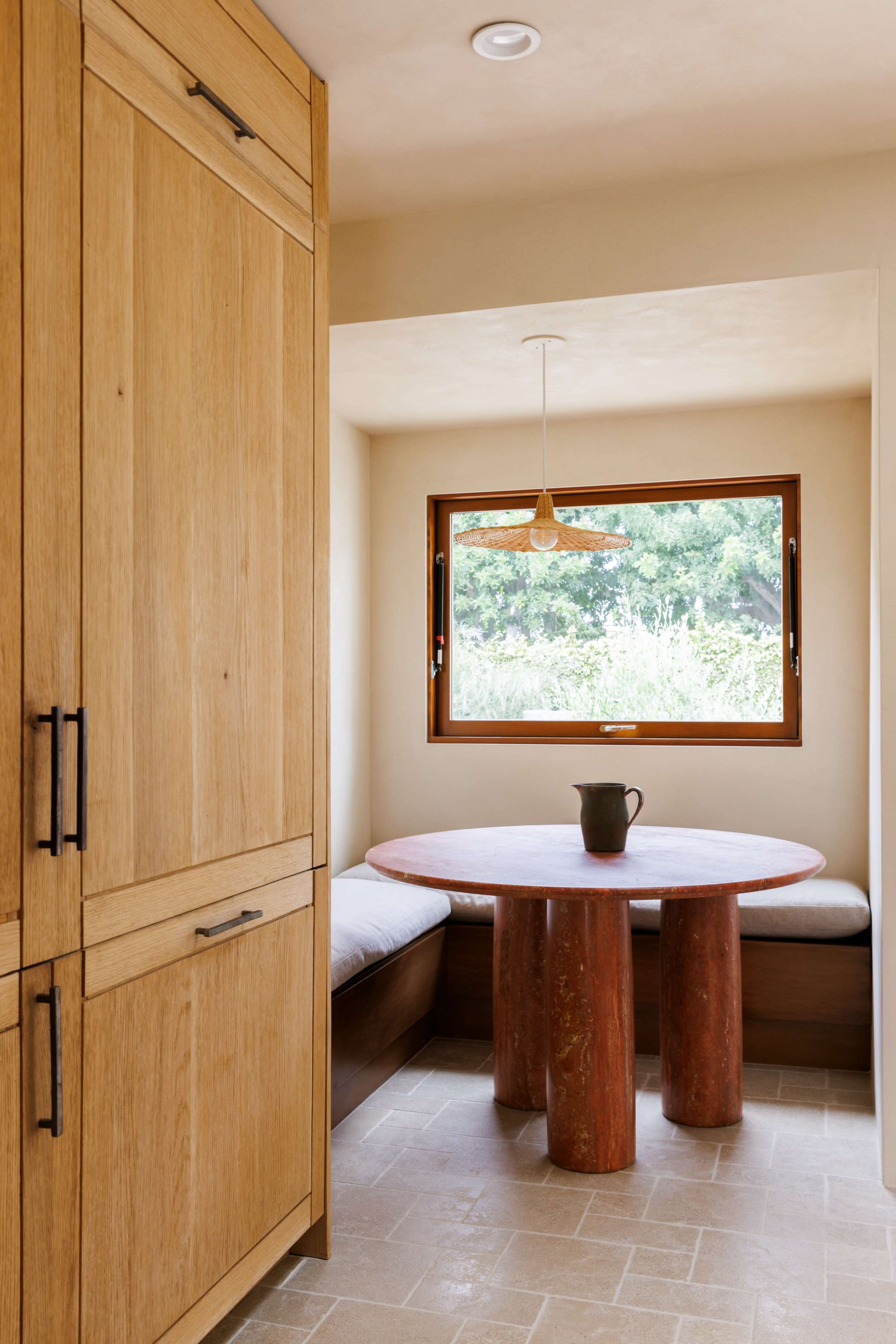 The image shows a small dining area featuring a round wooden table supported by cylindrical red pillars, with a bench along the wall and a window offering a view of greenery outside.