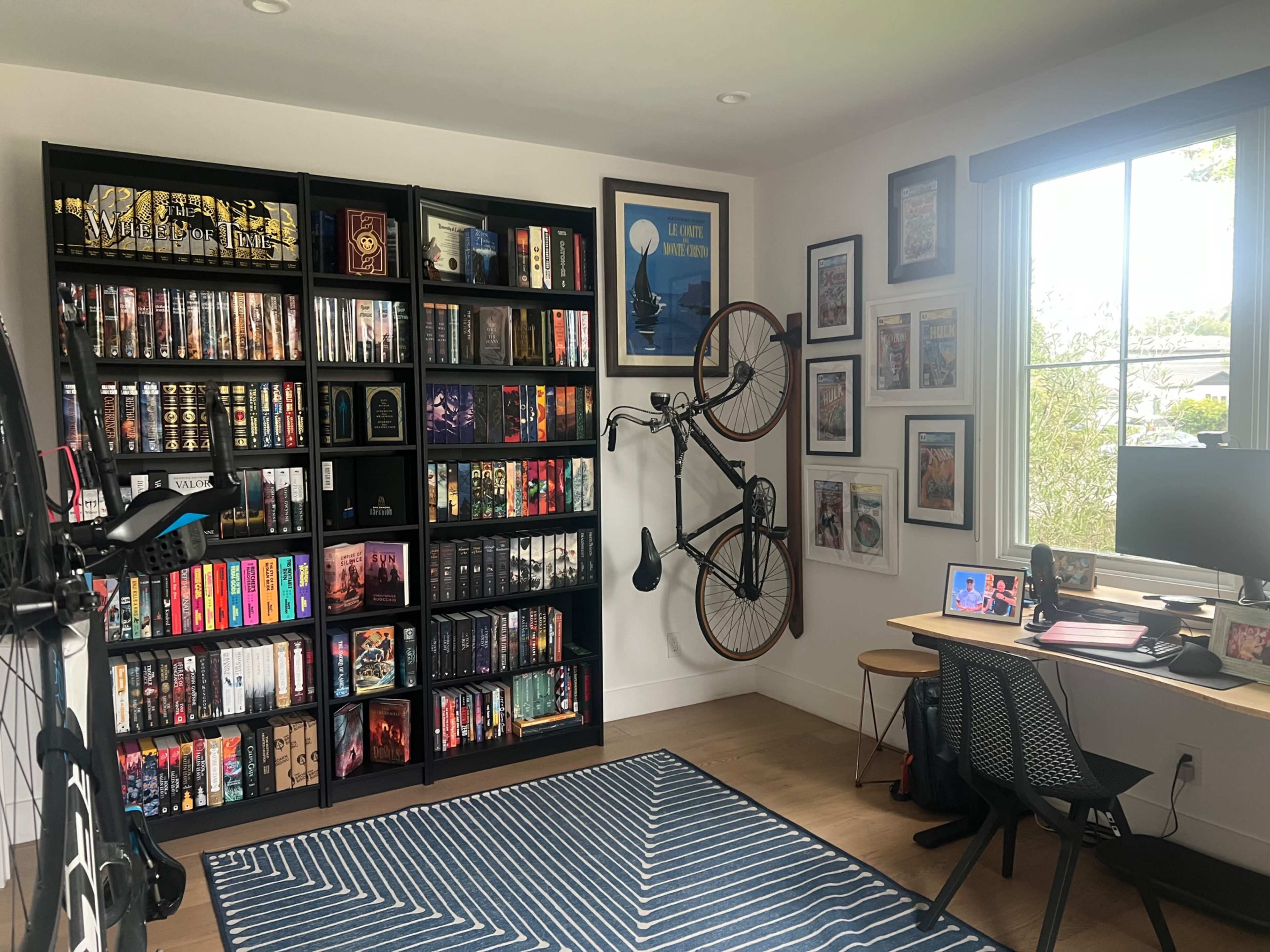 The image shows a home office featuring a large bookshelf filled with books, a mounted bicycle on the wall, and a desk with a computer and framed artwork.