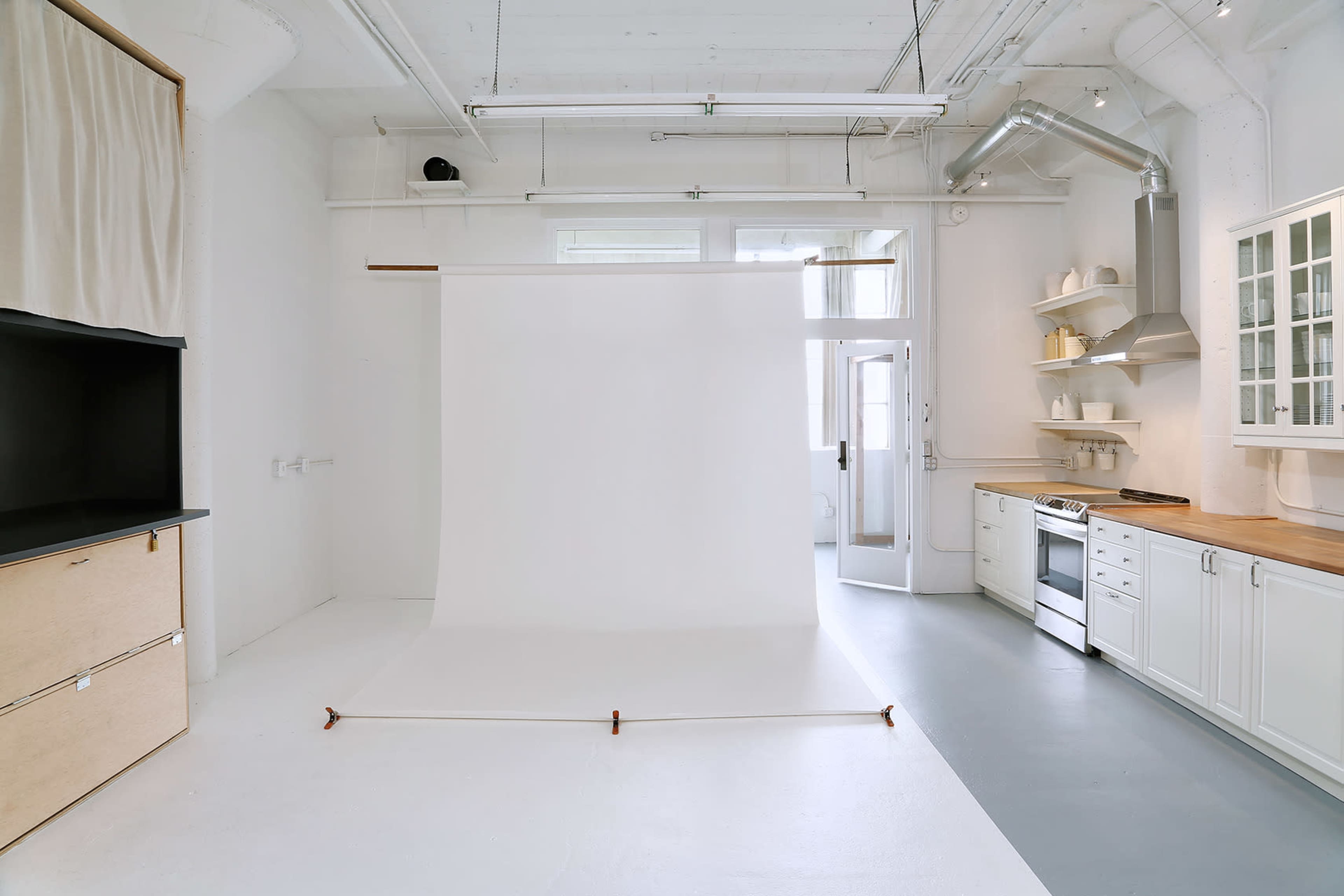 The image shows a bright, minimalist studio with a white backdrop, a small kitchen area, and open shelving.