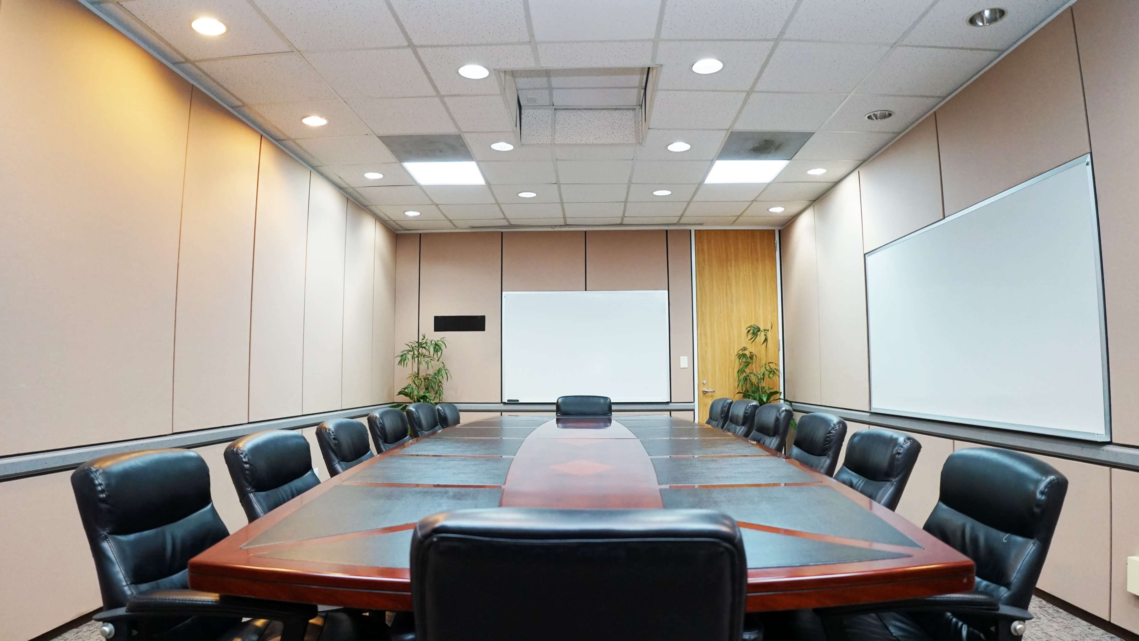 A conference room features a long rectangular table surrounded by black chairs, with whiteboards and plants along the walls.