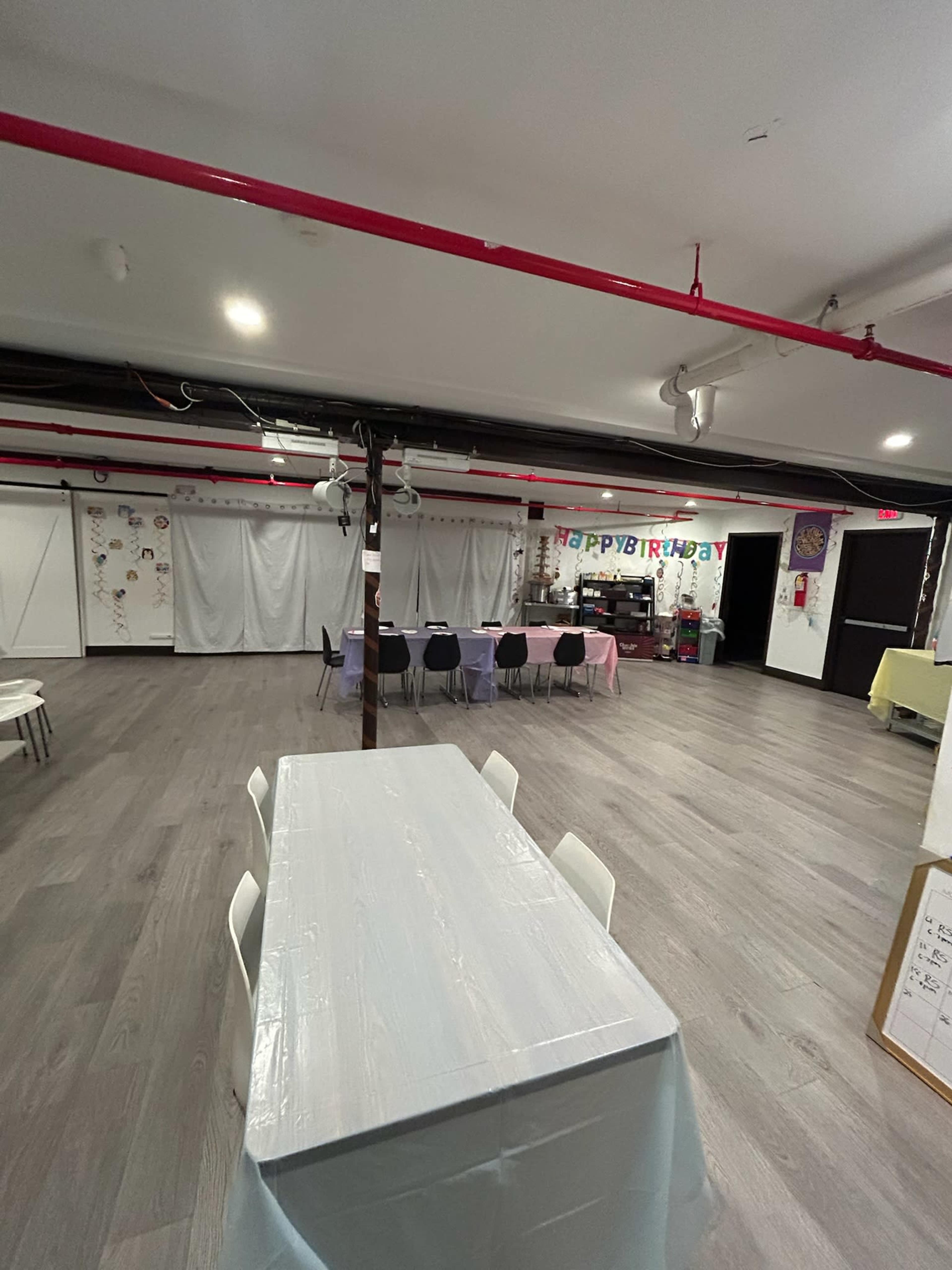 The image shows a spacious indoor party area with a long table covered in a light-colored cloth, surrounded by chairs, and decorated walls featuring birthday decorations.