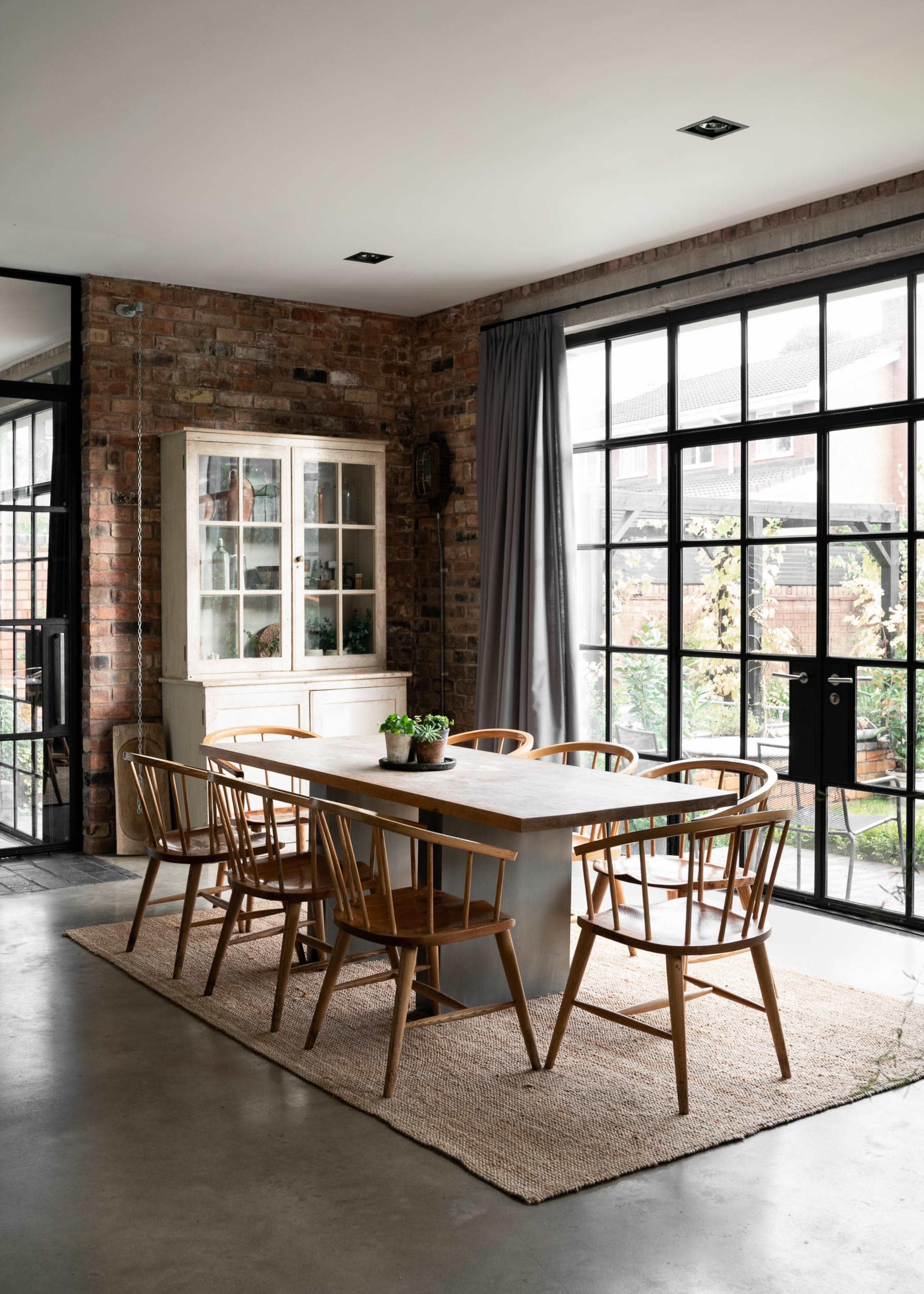 A modern dining area features a long table surrounded by wooden chairs, with large windows that overlook a garden.