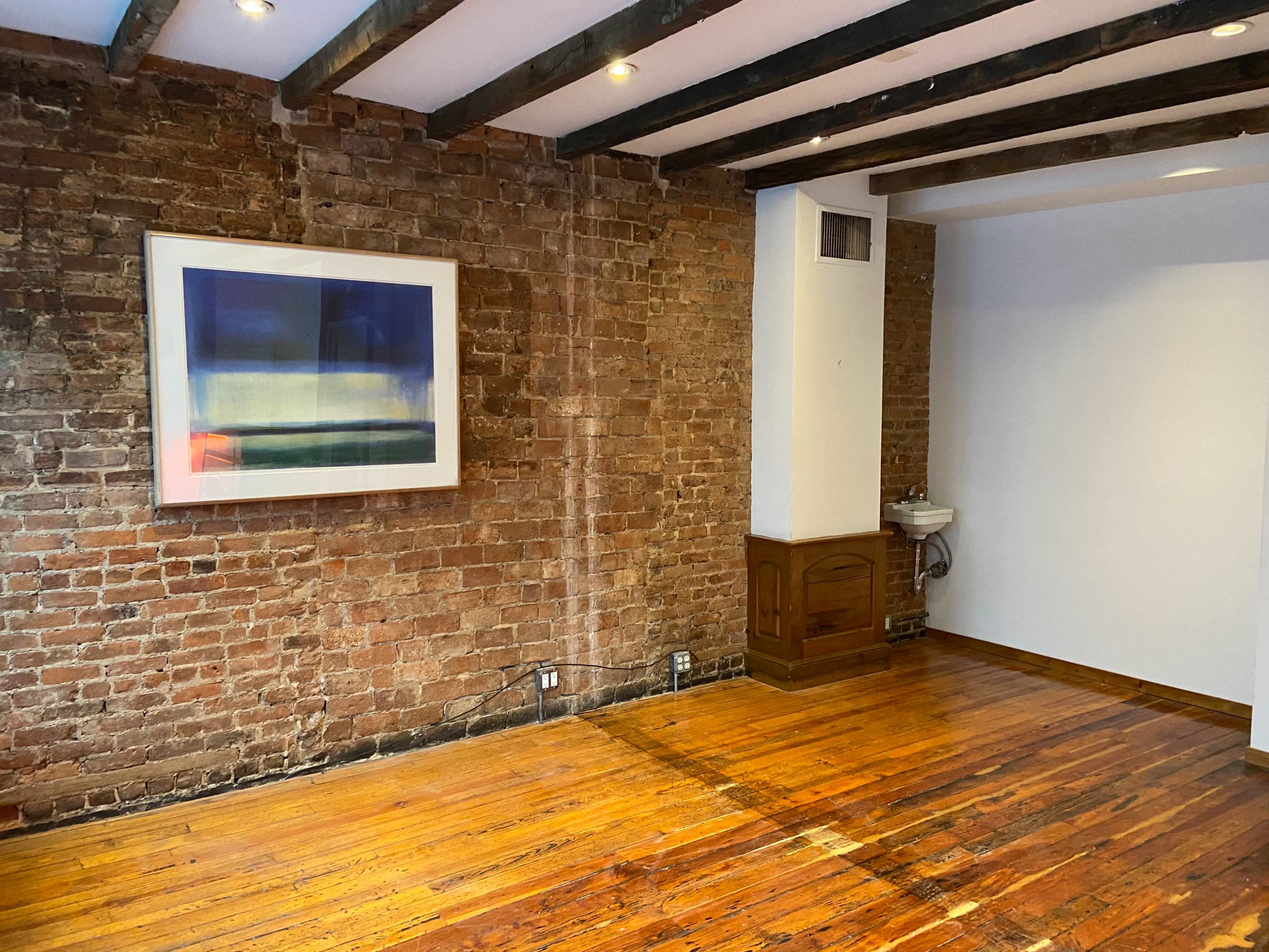 A room with exposed brick walls, wooden beam ceilings, and polished hardwood floors, featuring a large framed artwork on one wall and a small restroom area in the corner.