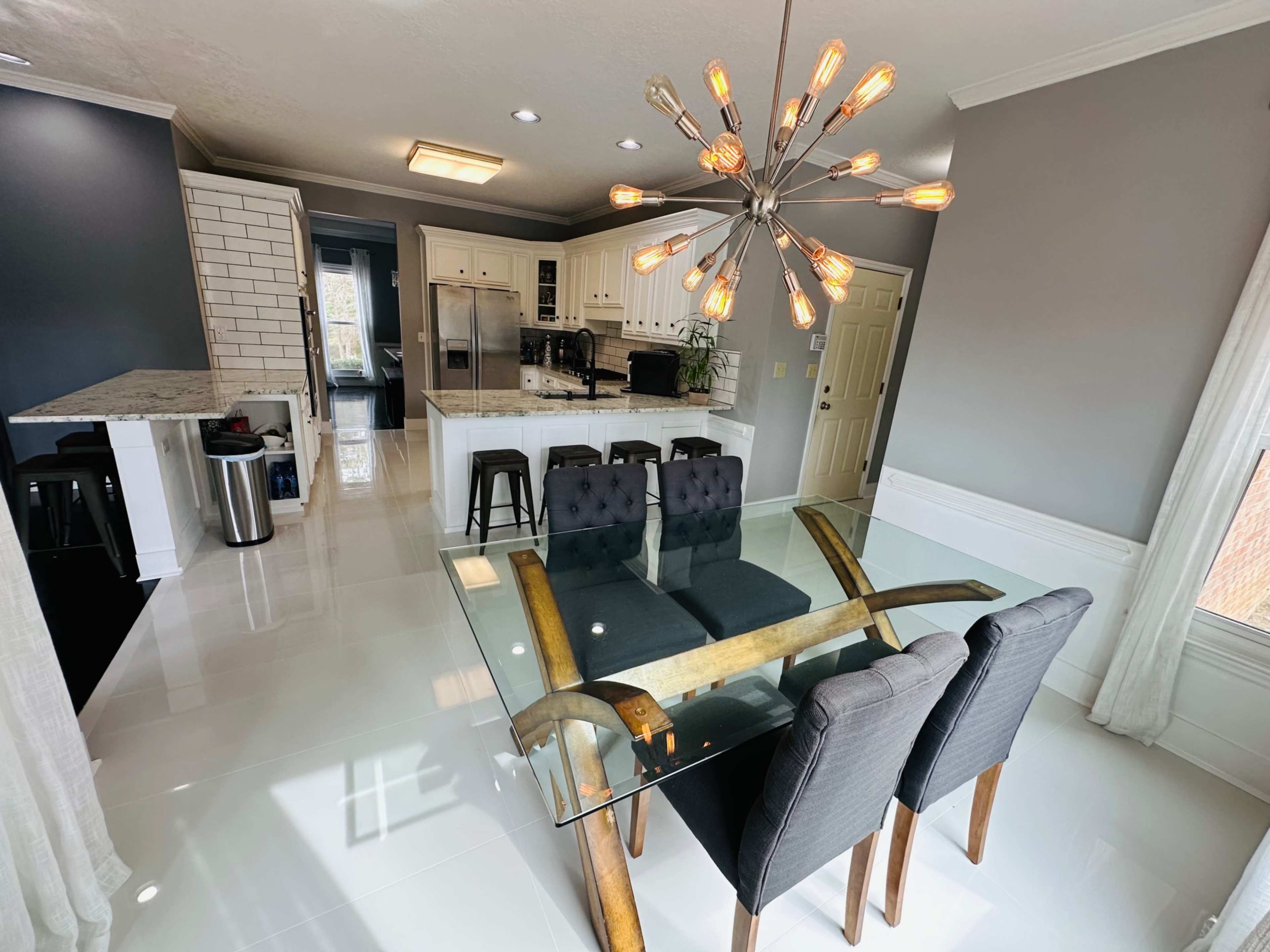 A modern kitchen and dining area features a glass-top table with four upholstered chairs, illuminated by a starburst chandelier.