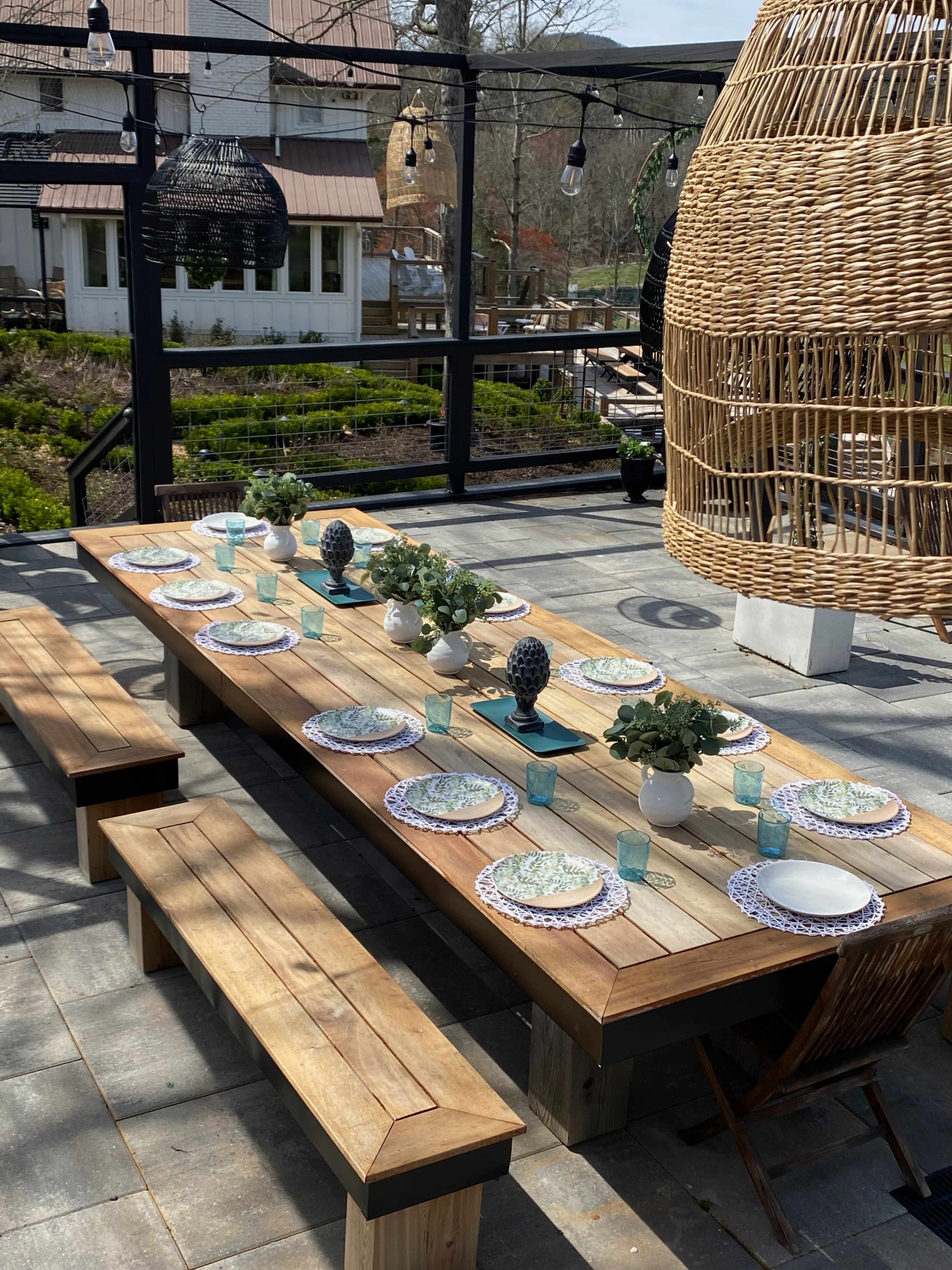 A long wooden table is set for a meal outdoors, featuring plates, glasses, and centerpieces, surrounded by benches and hanging decor.