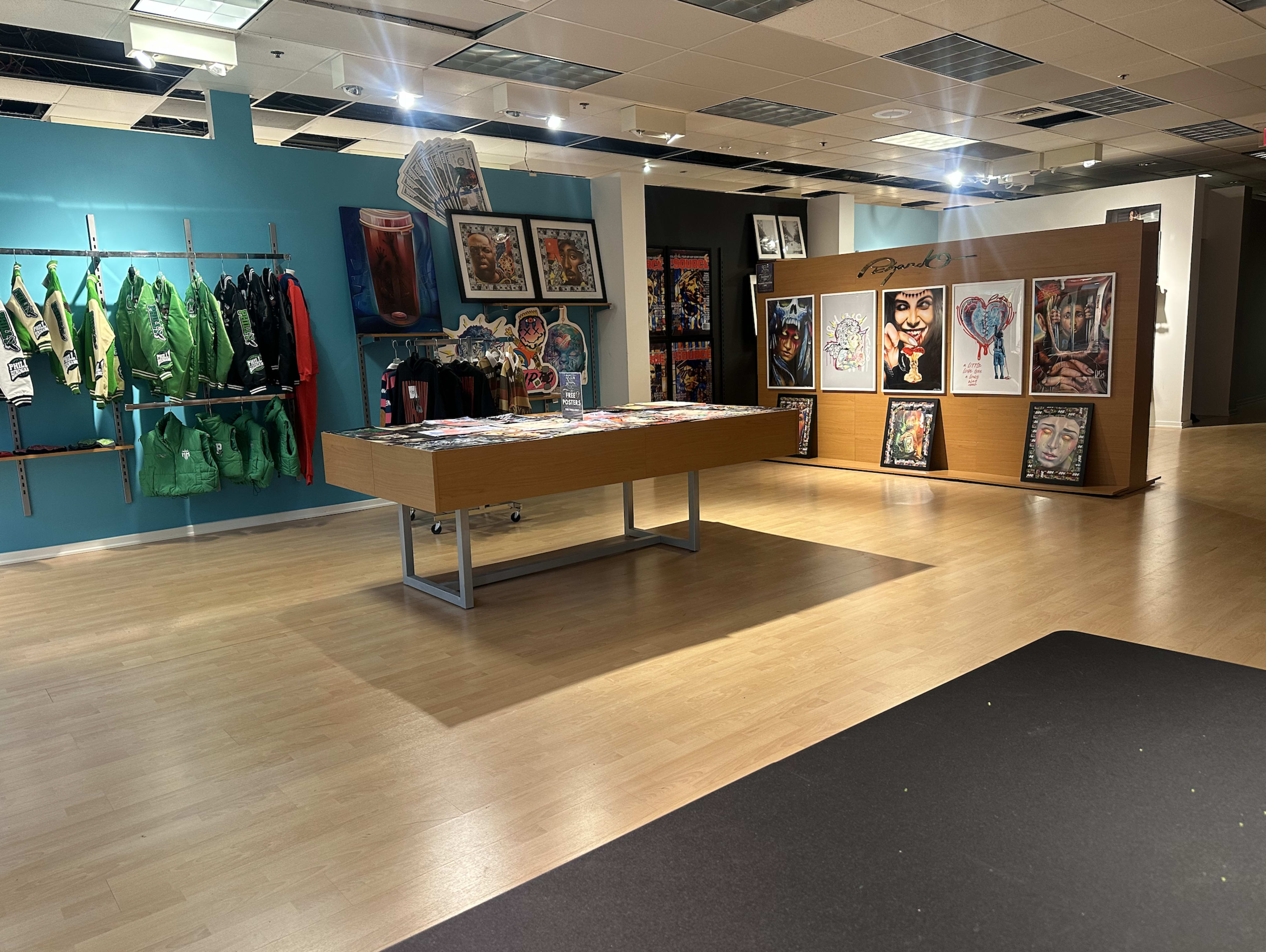 The image shows an art gallery with a wooden table displaying magazines, framed artworks on the walls, and clothing racks with green jackets in the background.