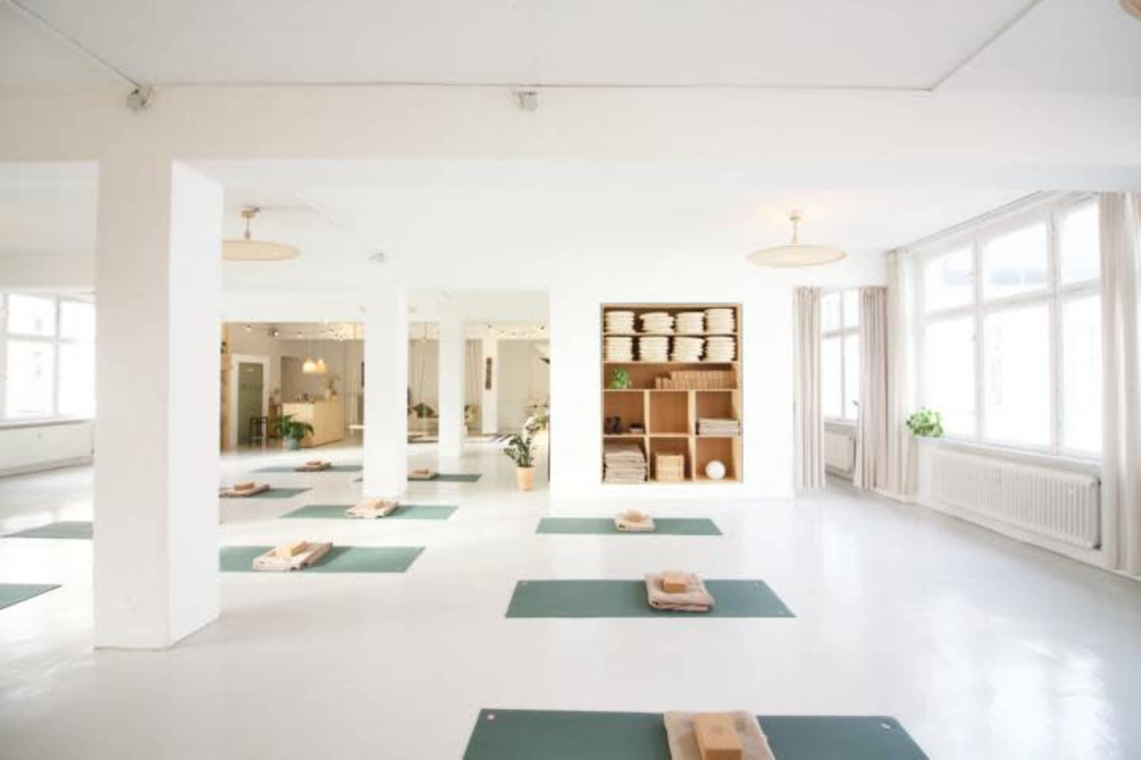 A spacious, well-lit yoga studio with green mats placed on a polished white floor and shelves containing supplies in the background.