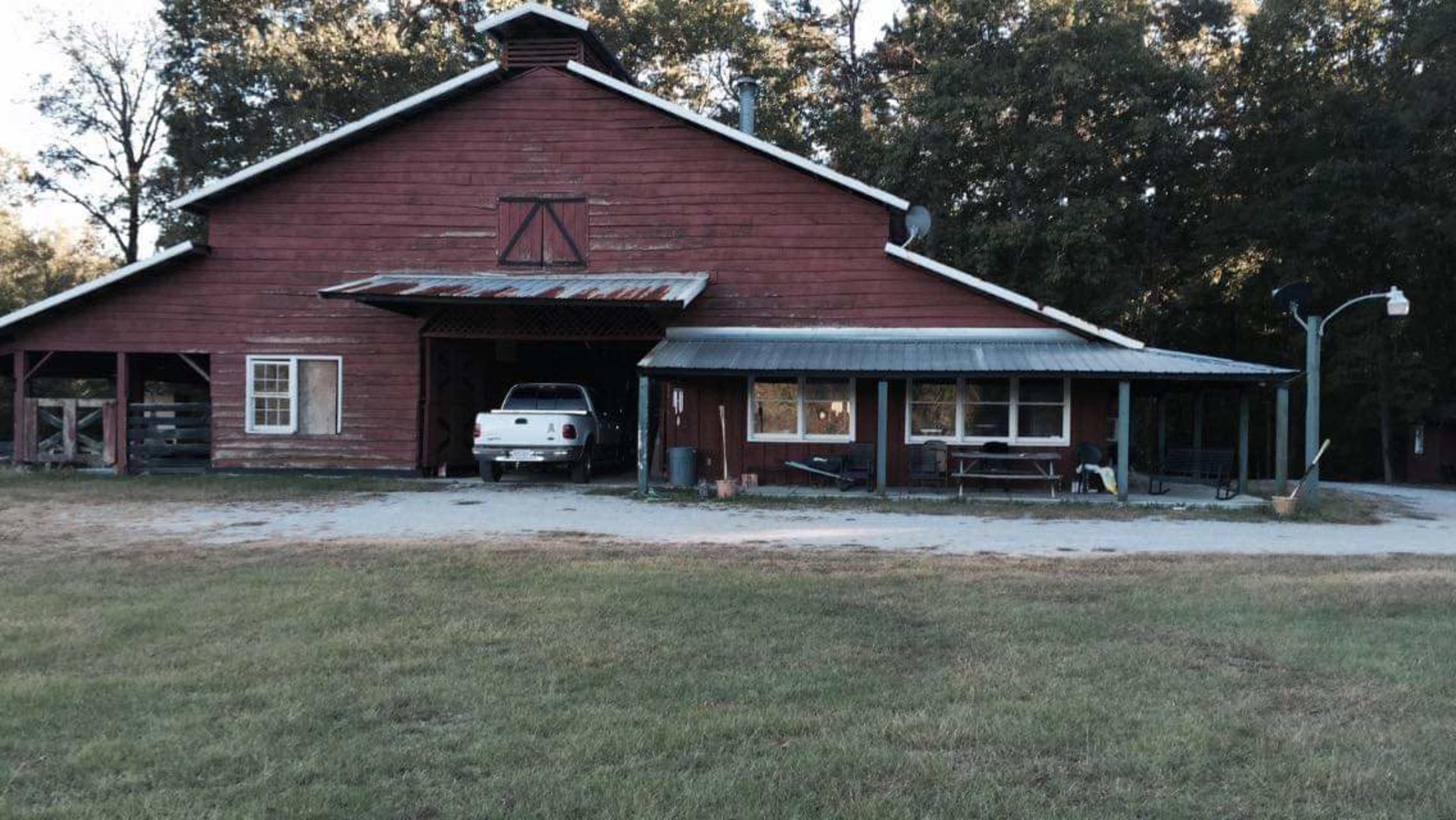 A red barn with a gray roof and a white truck parked in front is surrounded by a green lawn and trees.