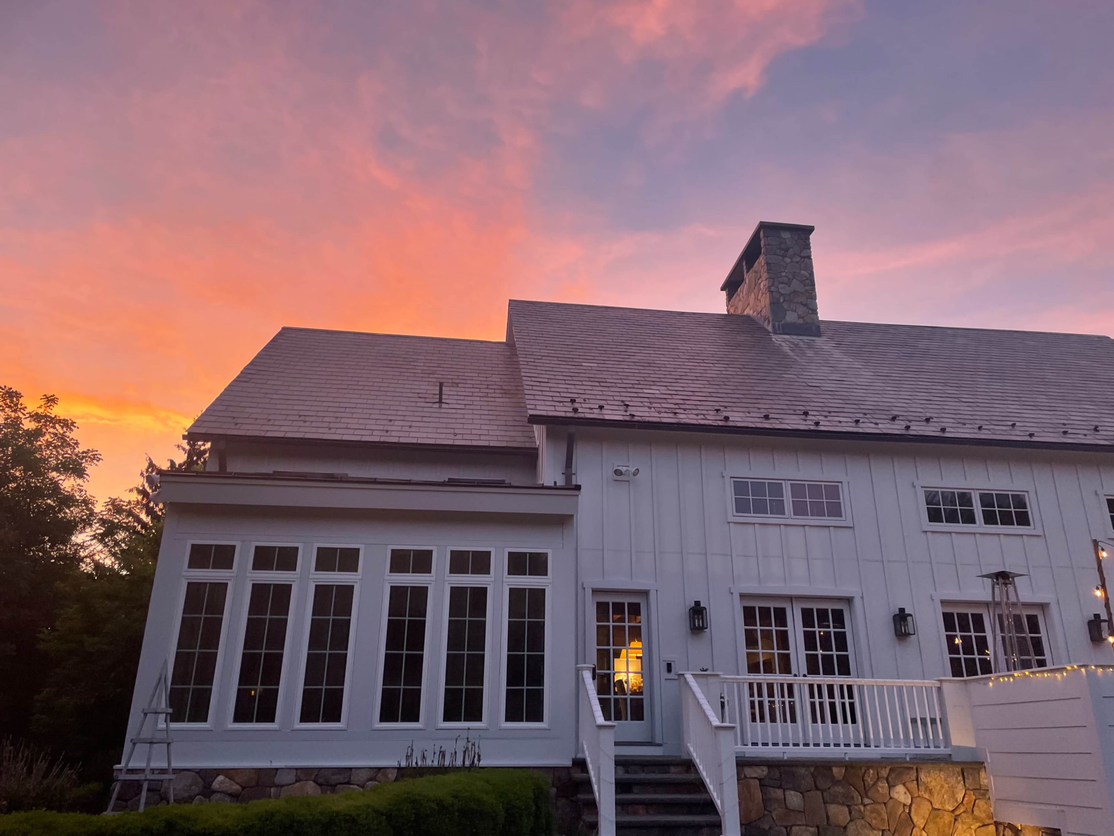 The image shows a white wooden house with large windows, a stone chimney, and a staircase leading up to a porch, set against a vibrant sunset sky.