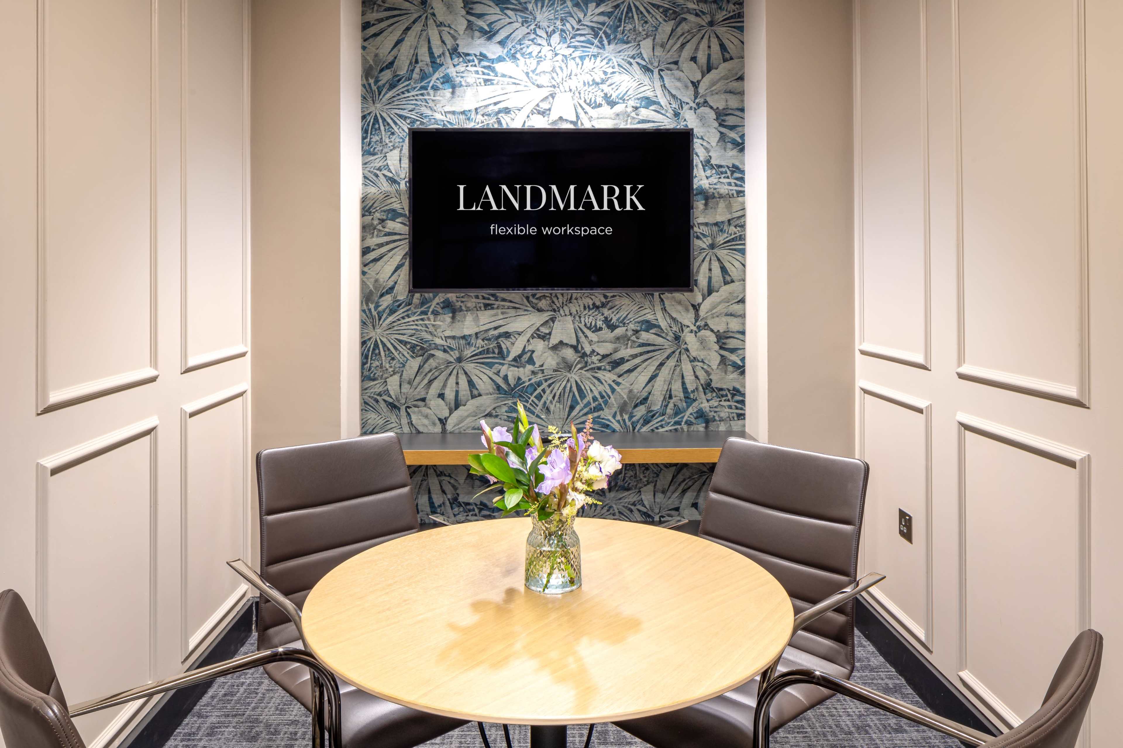 A small conference room features a round wooden table surrounded by four black chairs, with a floral arrangement in the center and a large screen displaying the "LANDMARK" logo on the wall.