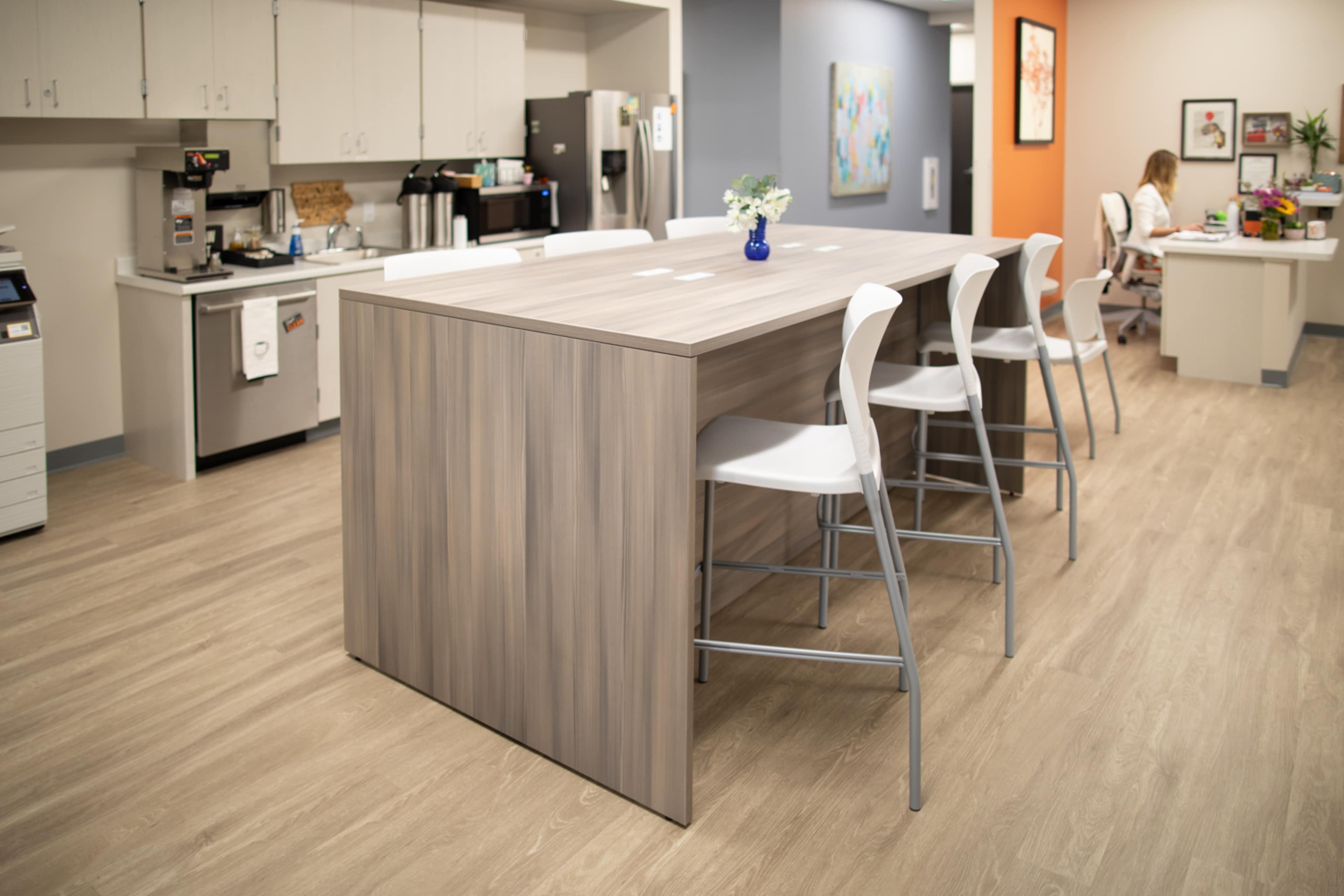 A modern kitchen area features a large wooden table surrounded by white chairs, with a small vase of flowers on the table and office supplies visible in the background.