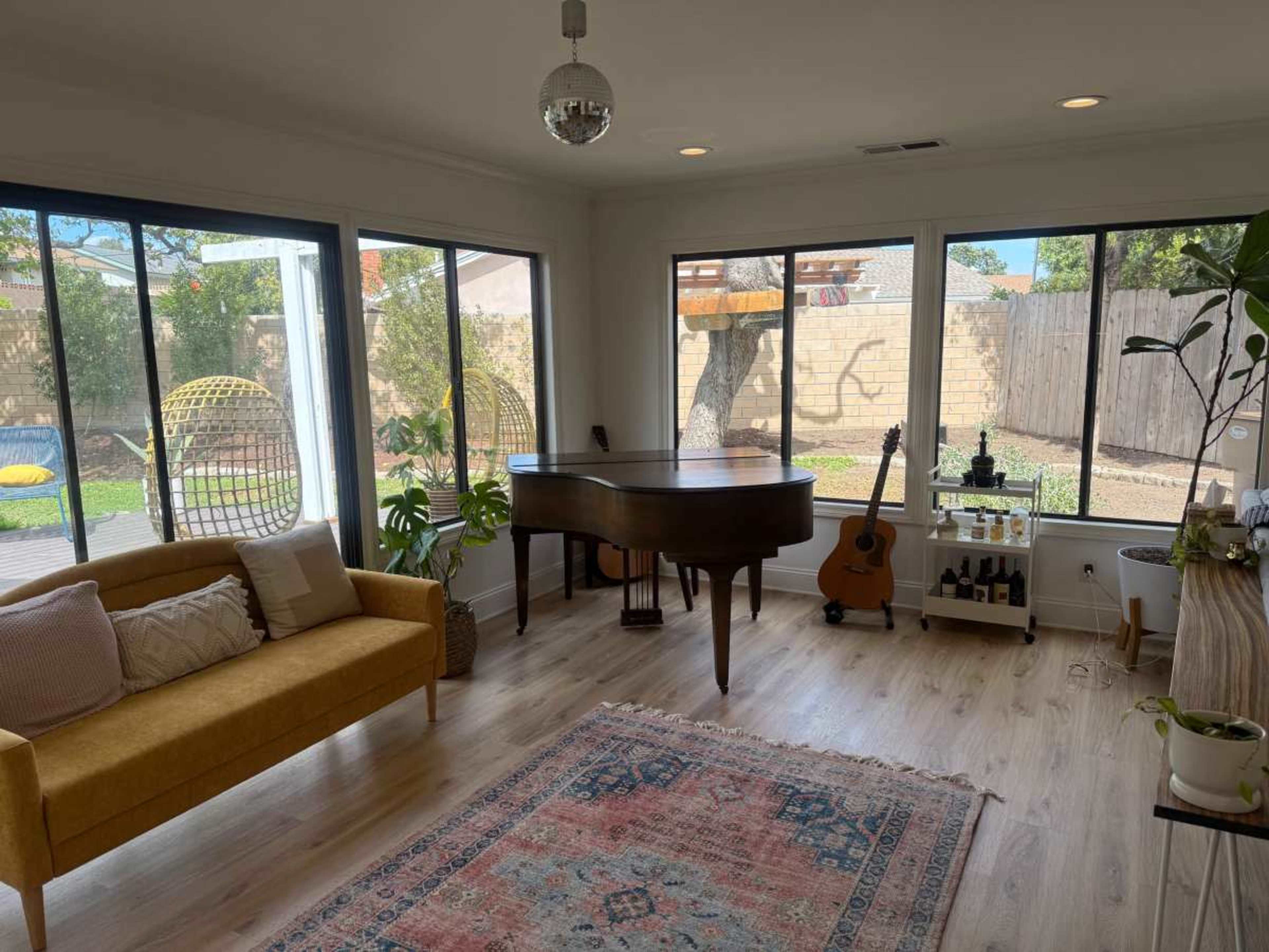A sunlit living room features a grand piano, a yellow sofa, and large windows revealing a backyard with greenery.