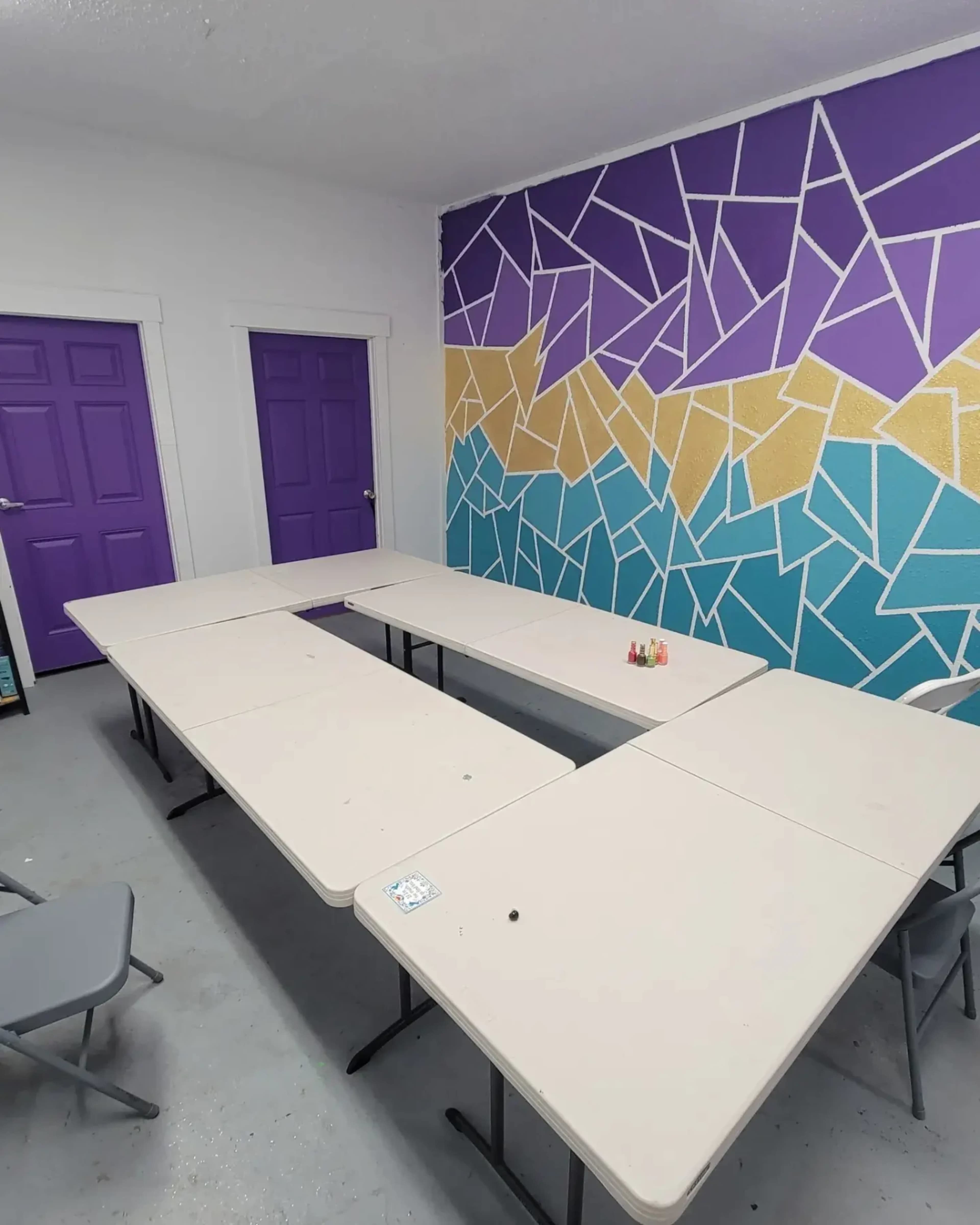 The image depicts a room with two purple doors and a wall featuring a colorful geometric mural, arranged with several tables forming a U-shape.