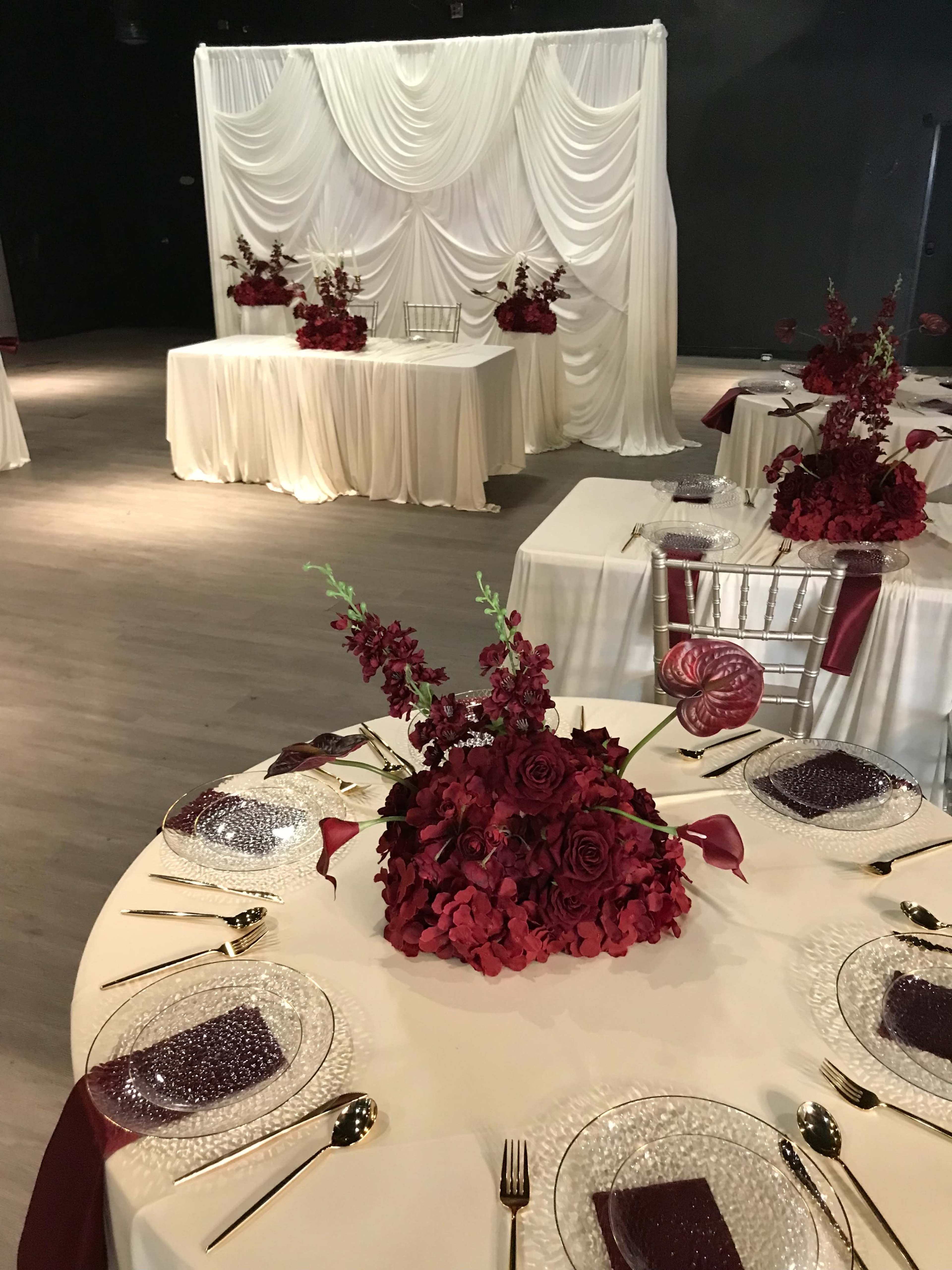 The image shows a banquet hall decorated with white draped fabric, tables set with elegant floral centerpieces in shades of red, and gold cutlery arranged on glass dinnerware.